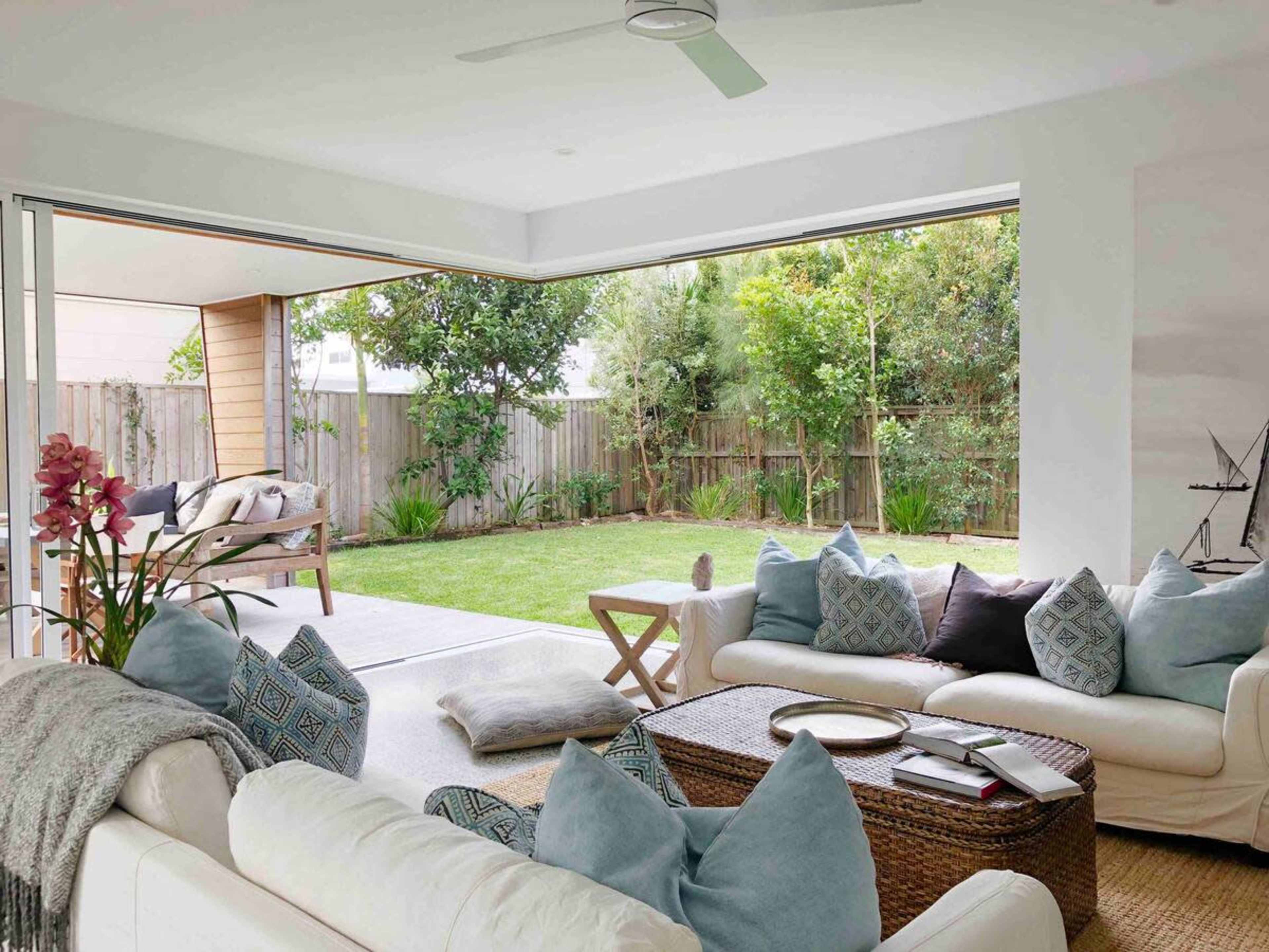 A spacious living room featuring a white sofa with decorative pillows and a coffee table, opening to a green backyard through large sliding glass doors.