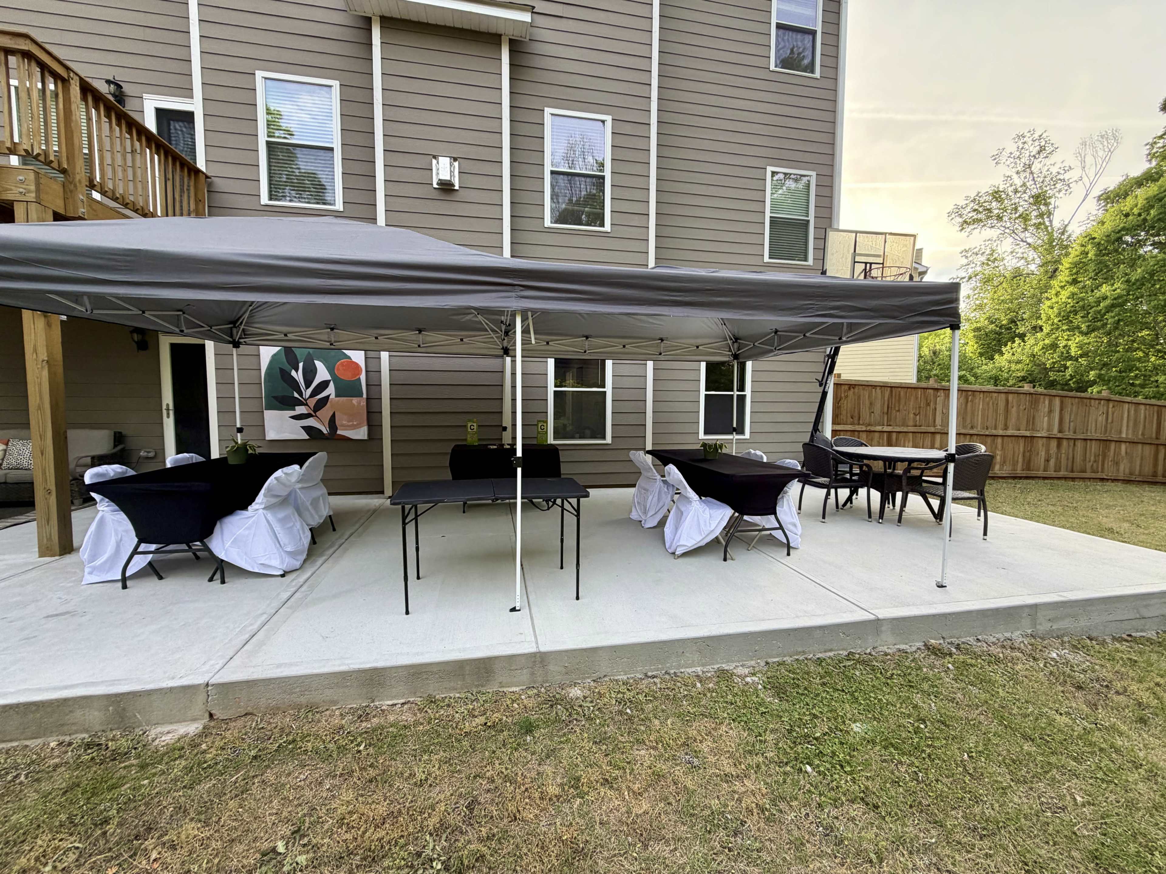 The image shows a concrete patio area with a large gray canopy, set up with several chairs and tables beneath it, adjacent to a two-story house with a wooden deck.