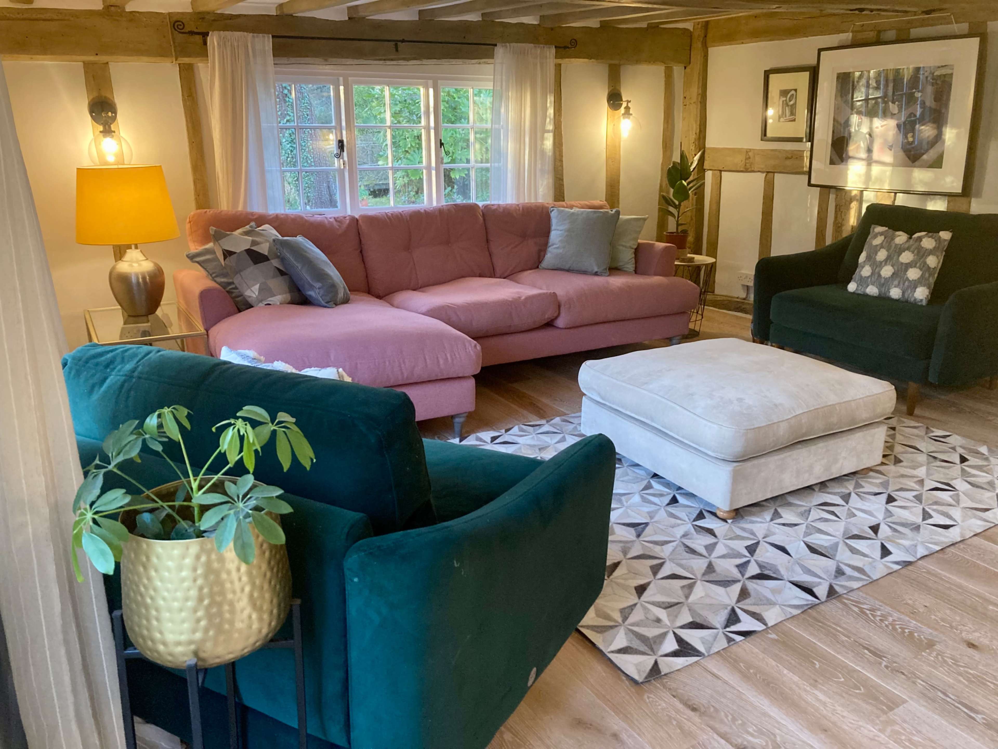 A cozy living room features a pink sectional sofa, a green armchair, a beige ottoman, and a geometric patterned rug under soft lighting.