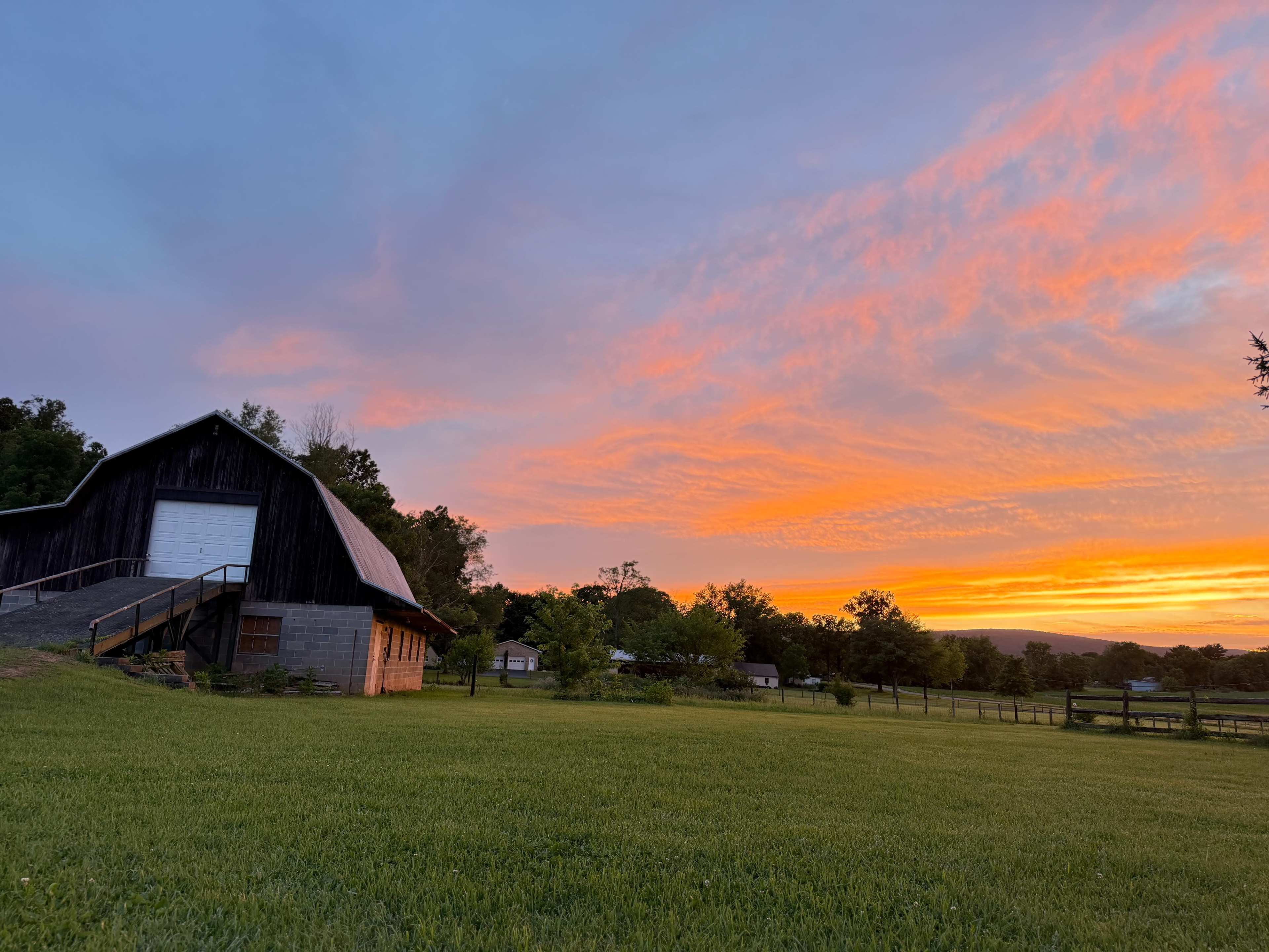 A barn stands in a grassy field under a vibrant sunset sky filled with clouds.