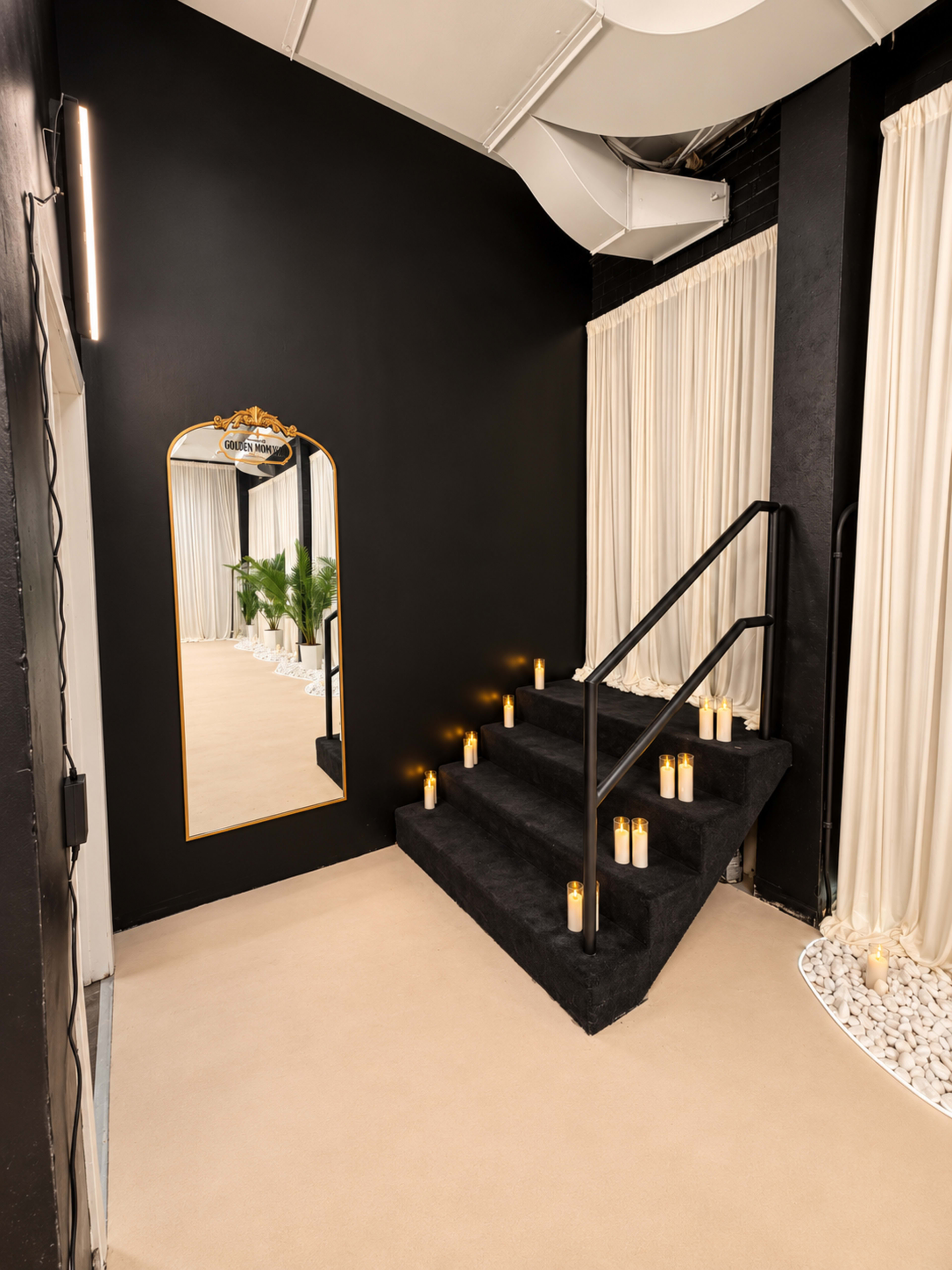 A staircase with black carpeting leads up to a mirror and is flanked by candles, creating a dramatic entryway adorned with white curtains and greenery.