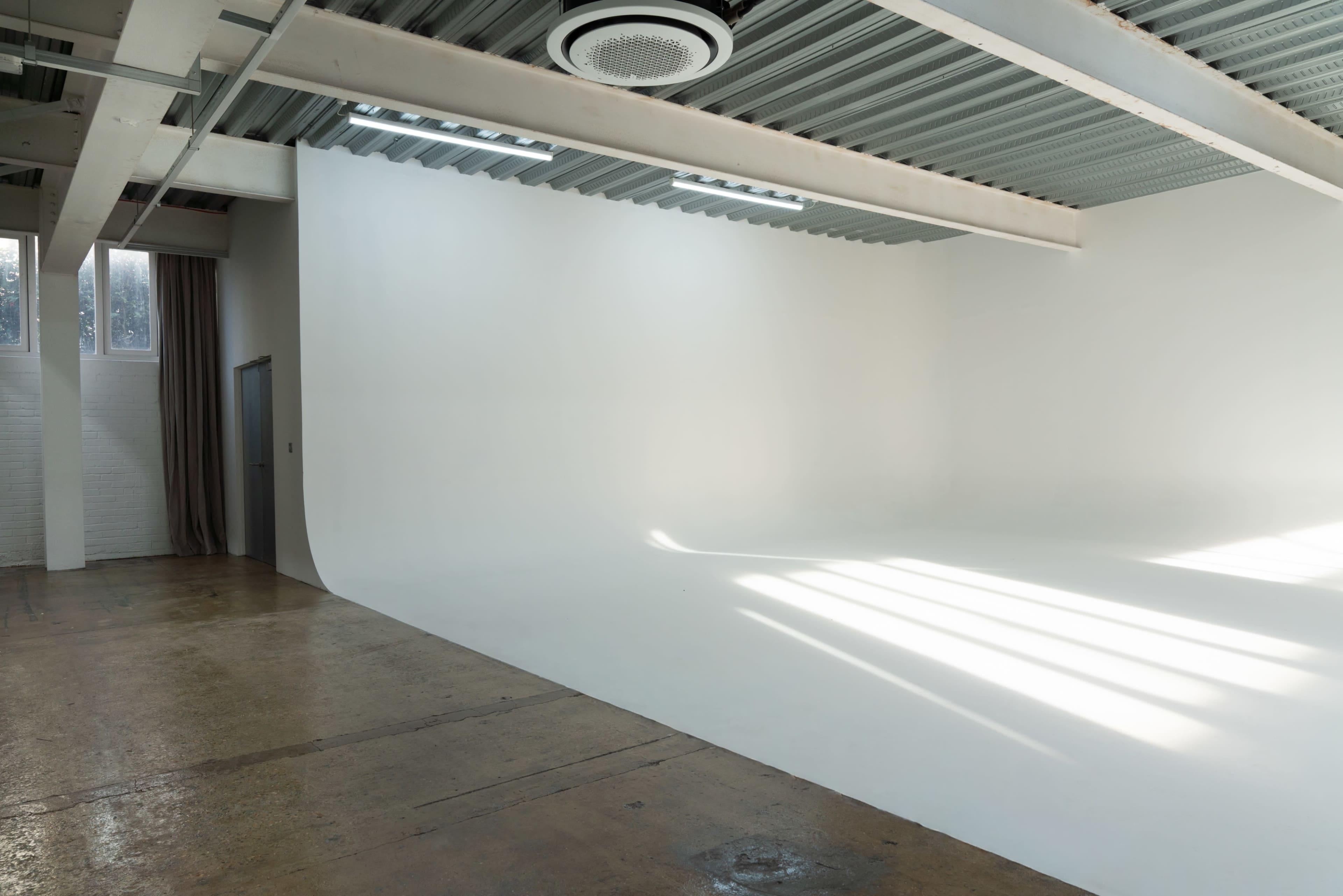 The image shows a spacious, white-walled studio with a seamless backdrop and natural light streaming in through large windows.