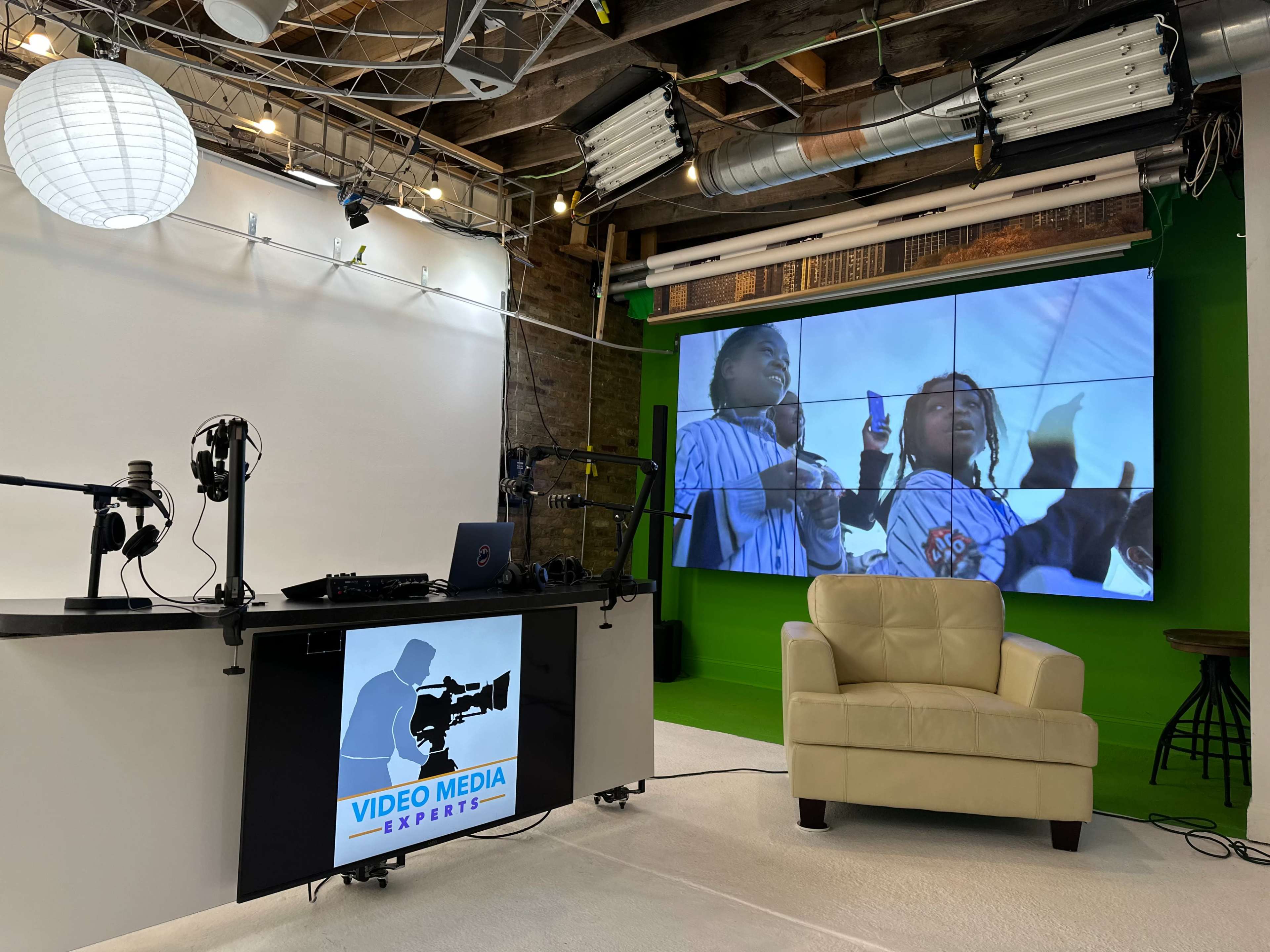 A media studio setup with a large screen displaying two children holding drinks, a cream-colored armchair, and microphones arranged on a black table.
