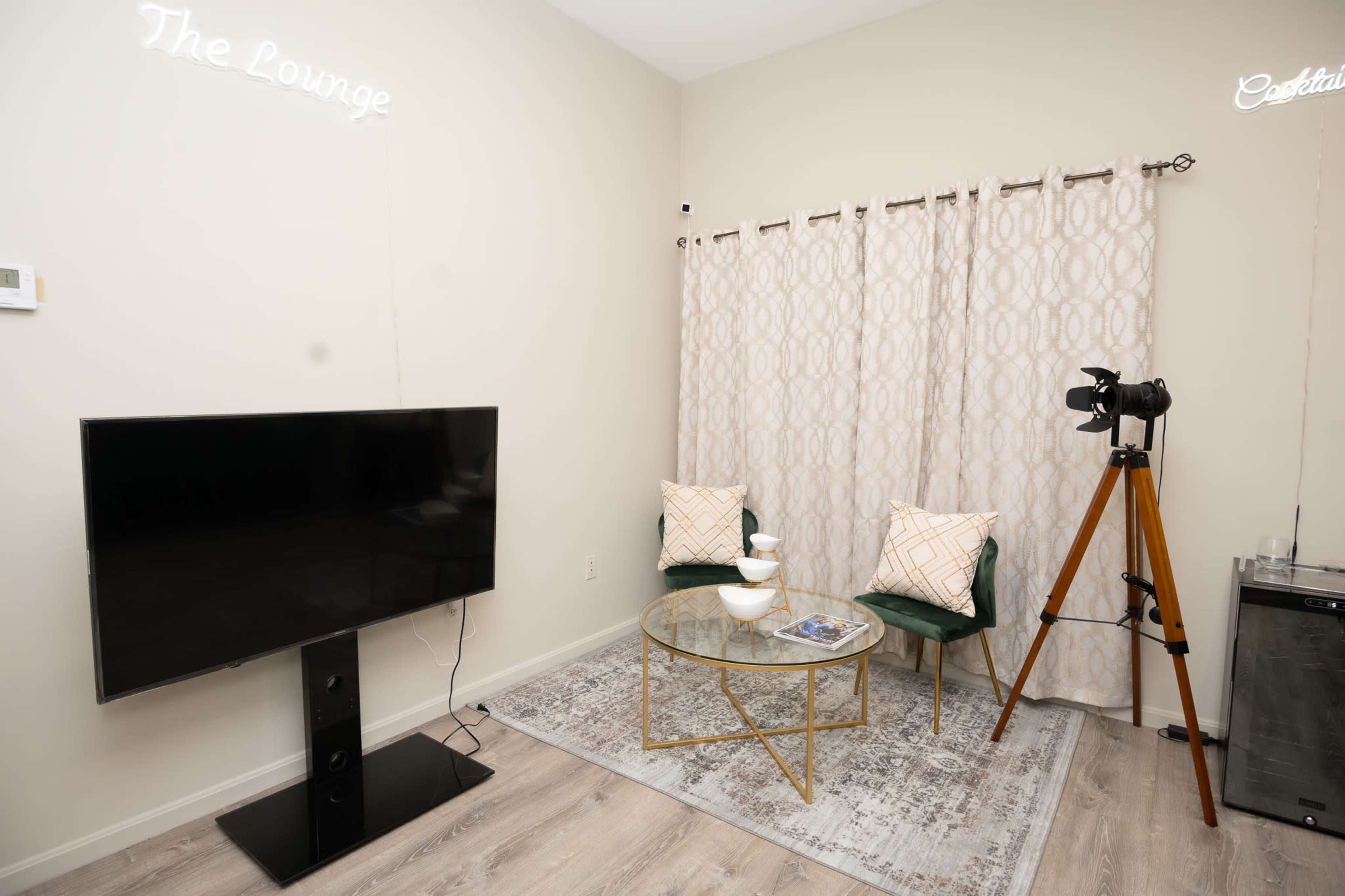 The image shows a small lounge area featuring a television, a glass coffee table, two chairs with decorative pillows, a window with curtains, and a camera on a tripod.