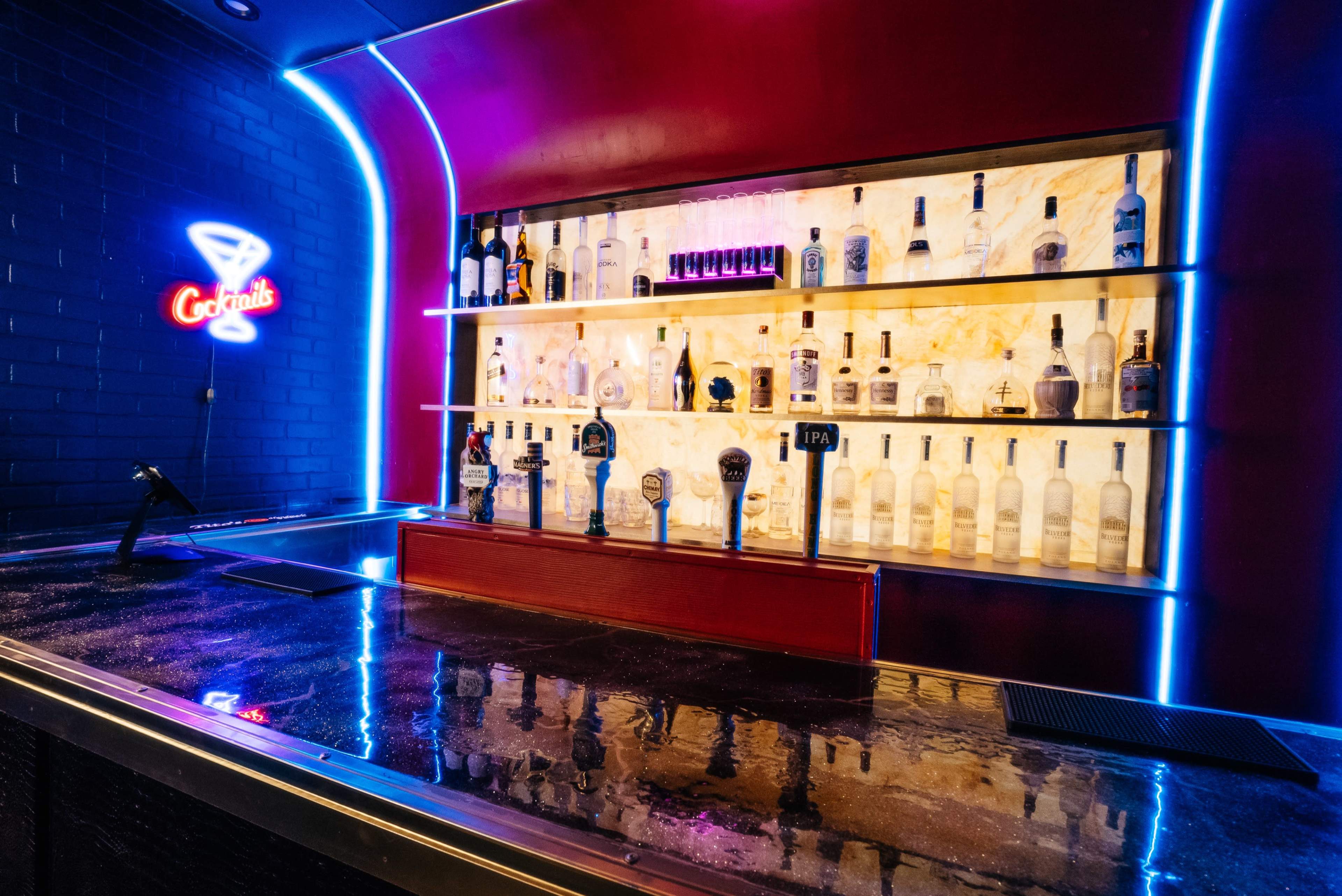 A well-lit bar with a variety of bottles displayed on shelves against a red backdrop, and a neon sign that reads "Cocktails."