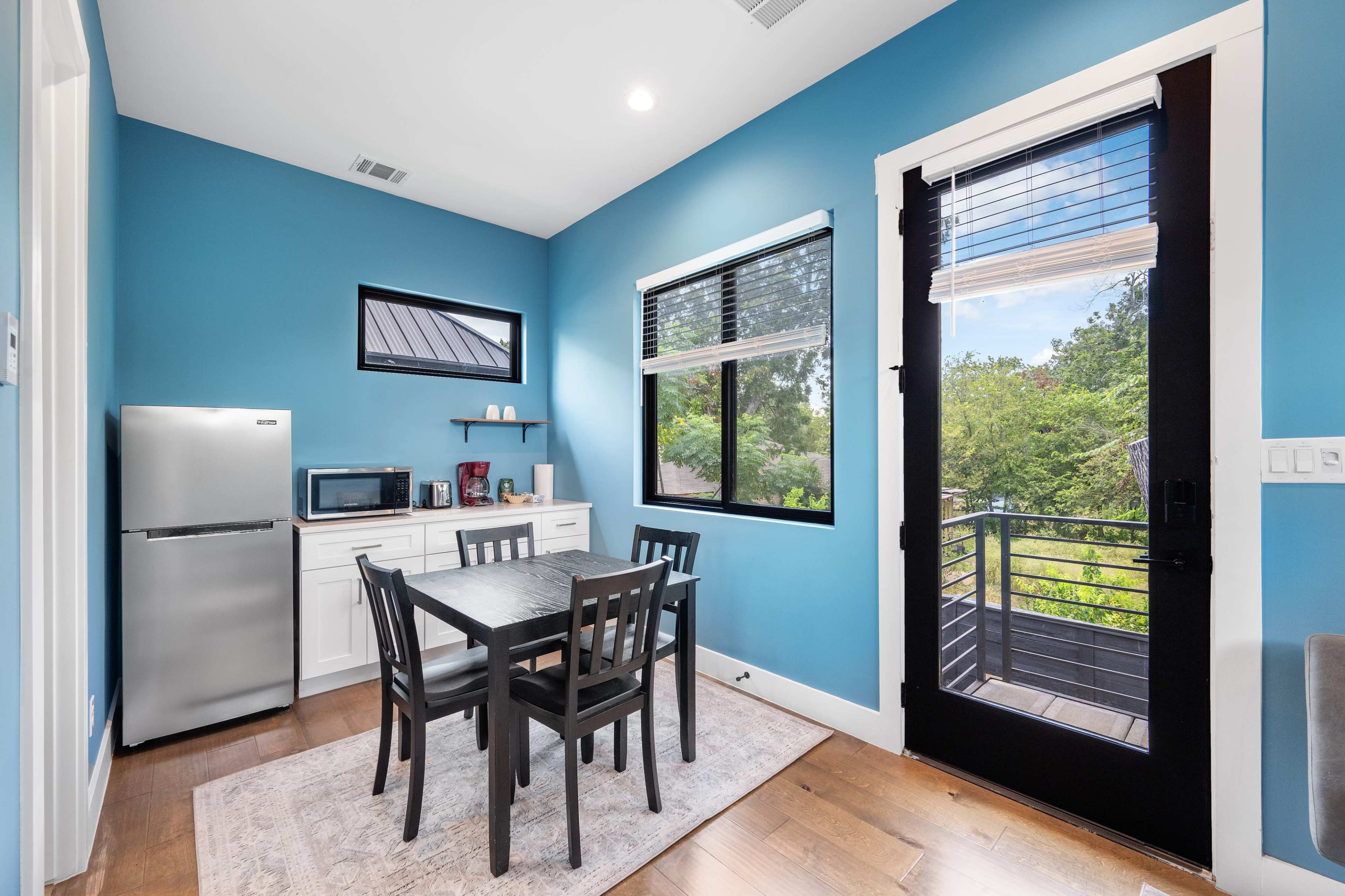 The image shows a small kitchen area with a table and chairs, a stainless steel refrigerator, and blue walls with large windows allowing natural light to enter.