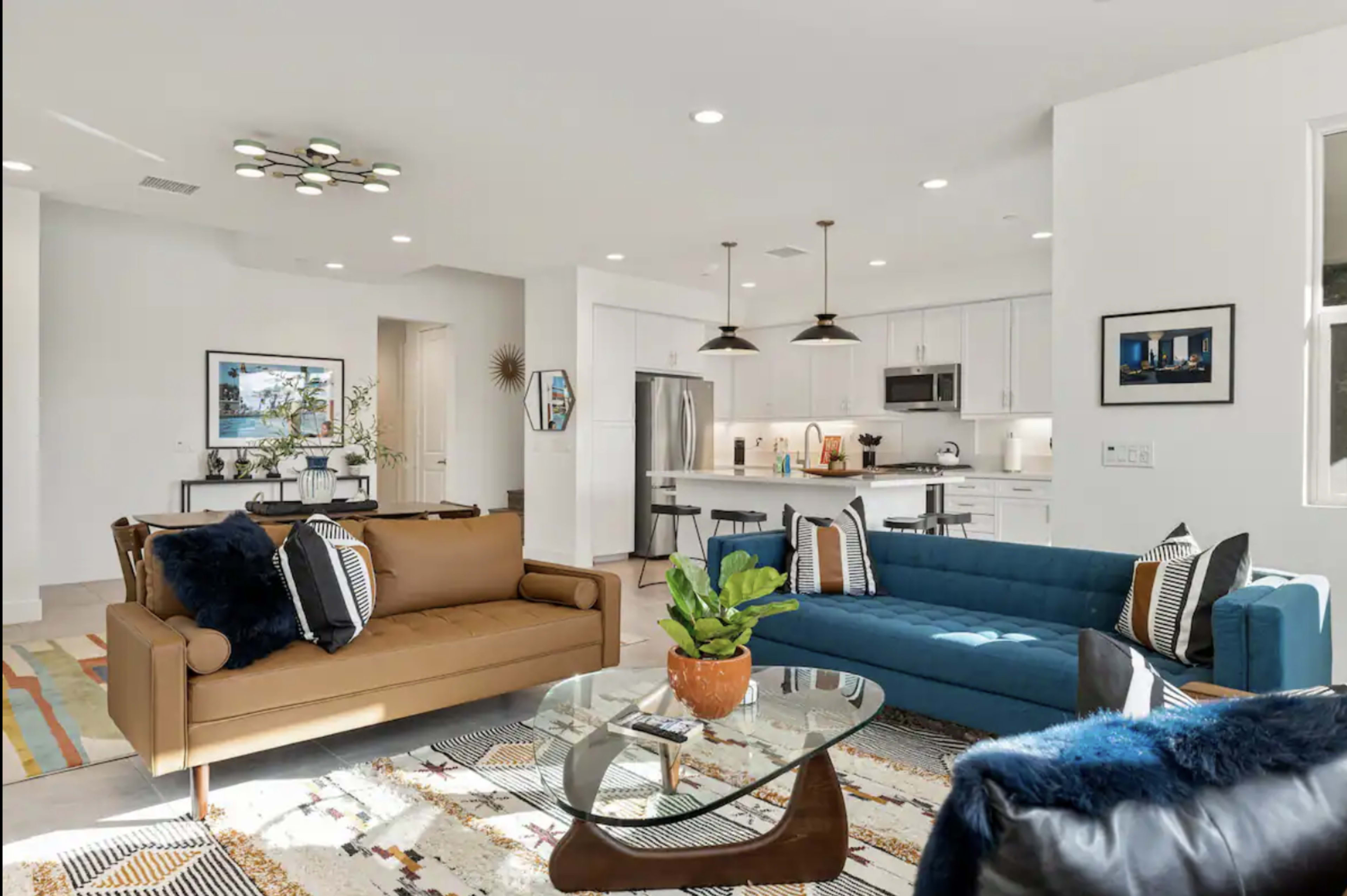 The living room features a brown sofa and a blue sofa arranged around a glass coffee table, with a colorful area rug beneath and a kitchen visible in the background.