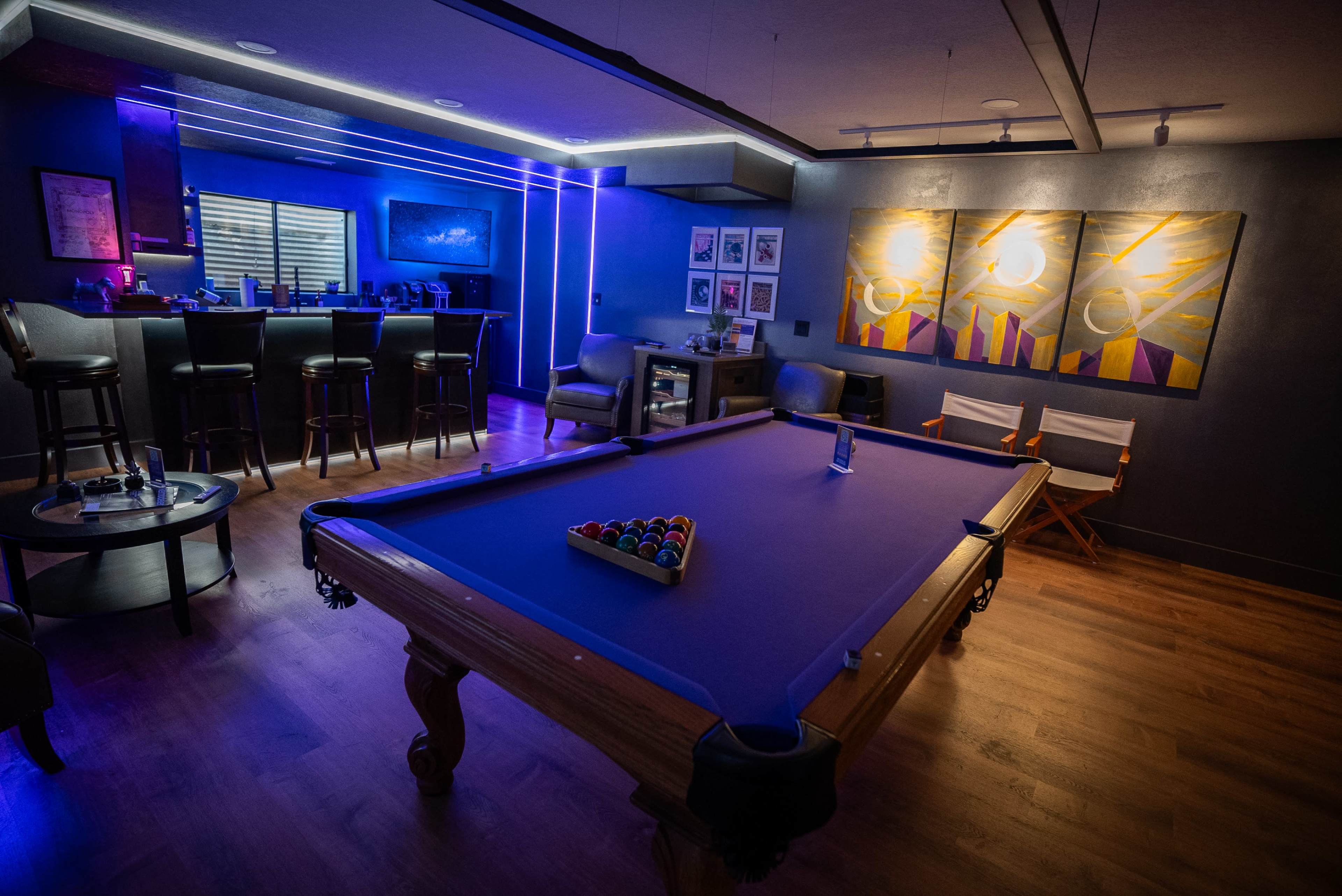 A billiards room features a purple pool table, surrounded by furniture, a bar area, and illuminated by ambient lighting.