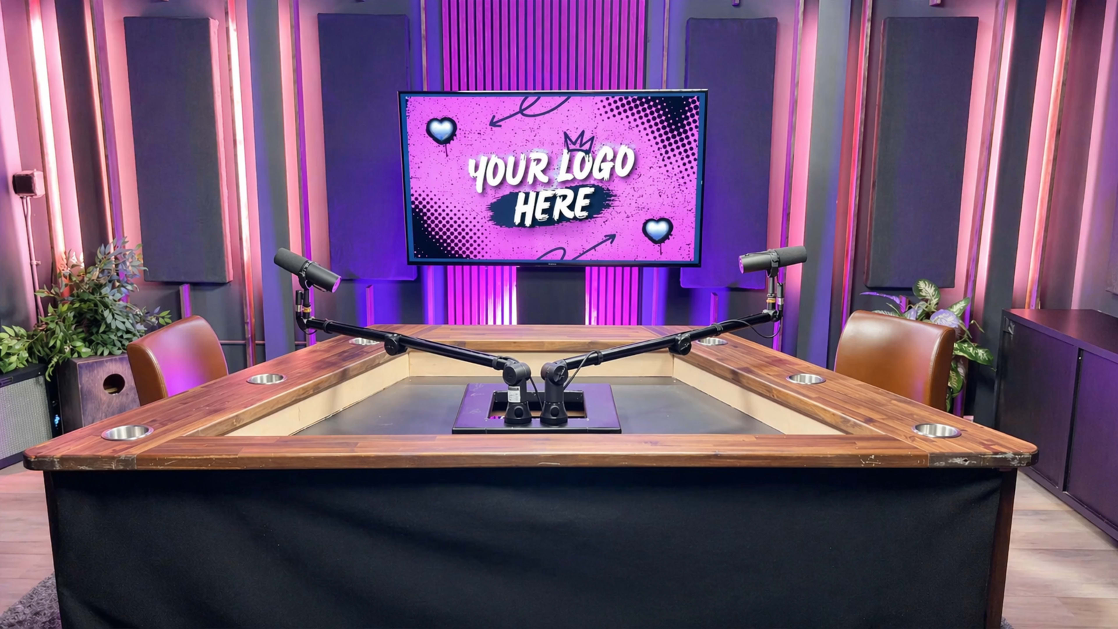 The image shows a podcast recording studio with a wooden table featuring two microphone stands, and a screen in the background displaying text that says "YOUR LOGO HERE" against a purple backdrop.