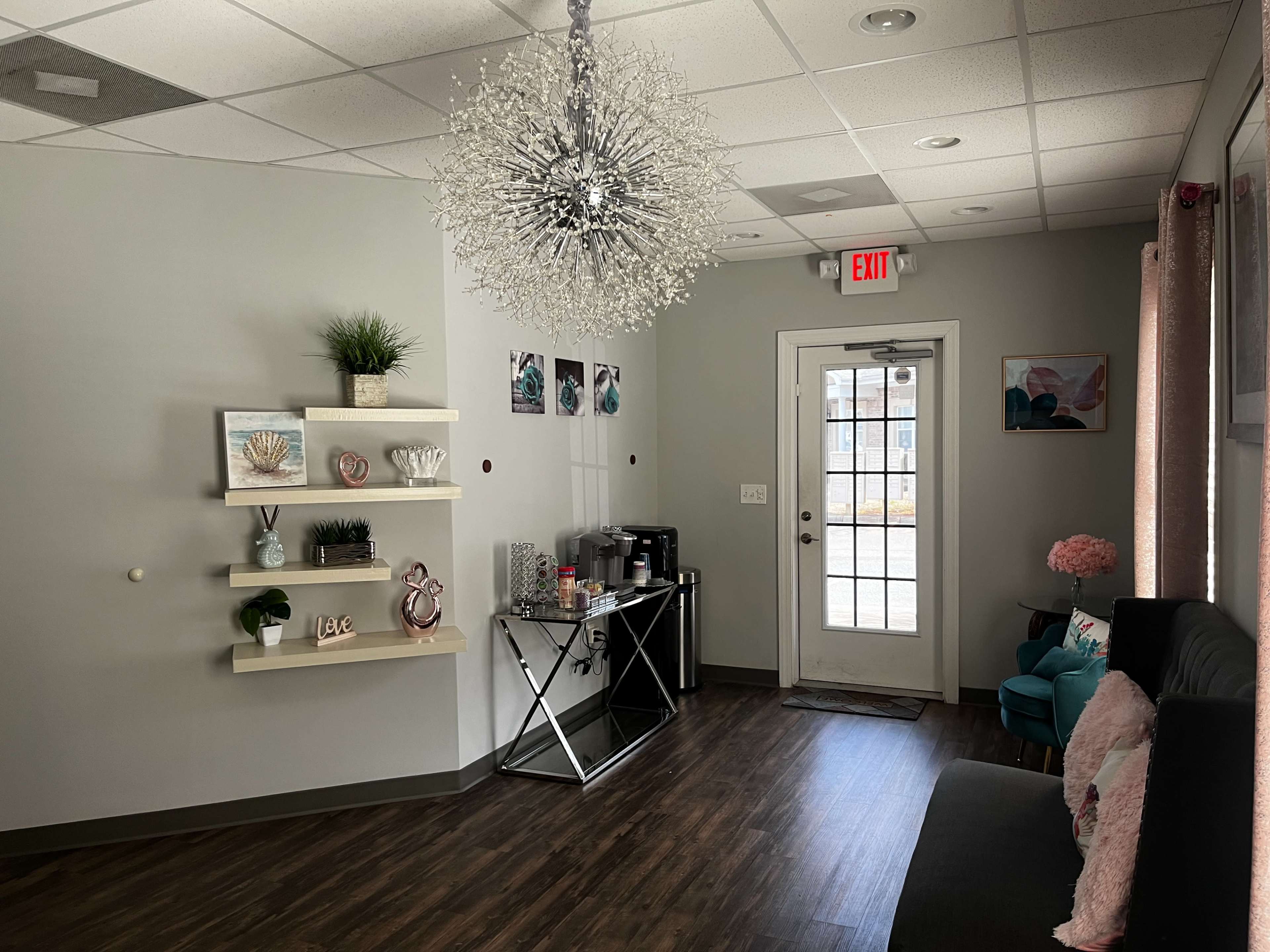 A modern reception area featuring a stylish chandelier, a wall-mounted shelf with decorative items, a sleek table with a coffee station, and a door leading outside.