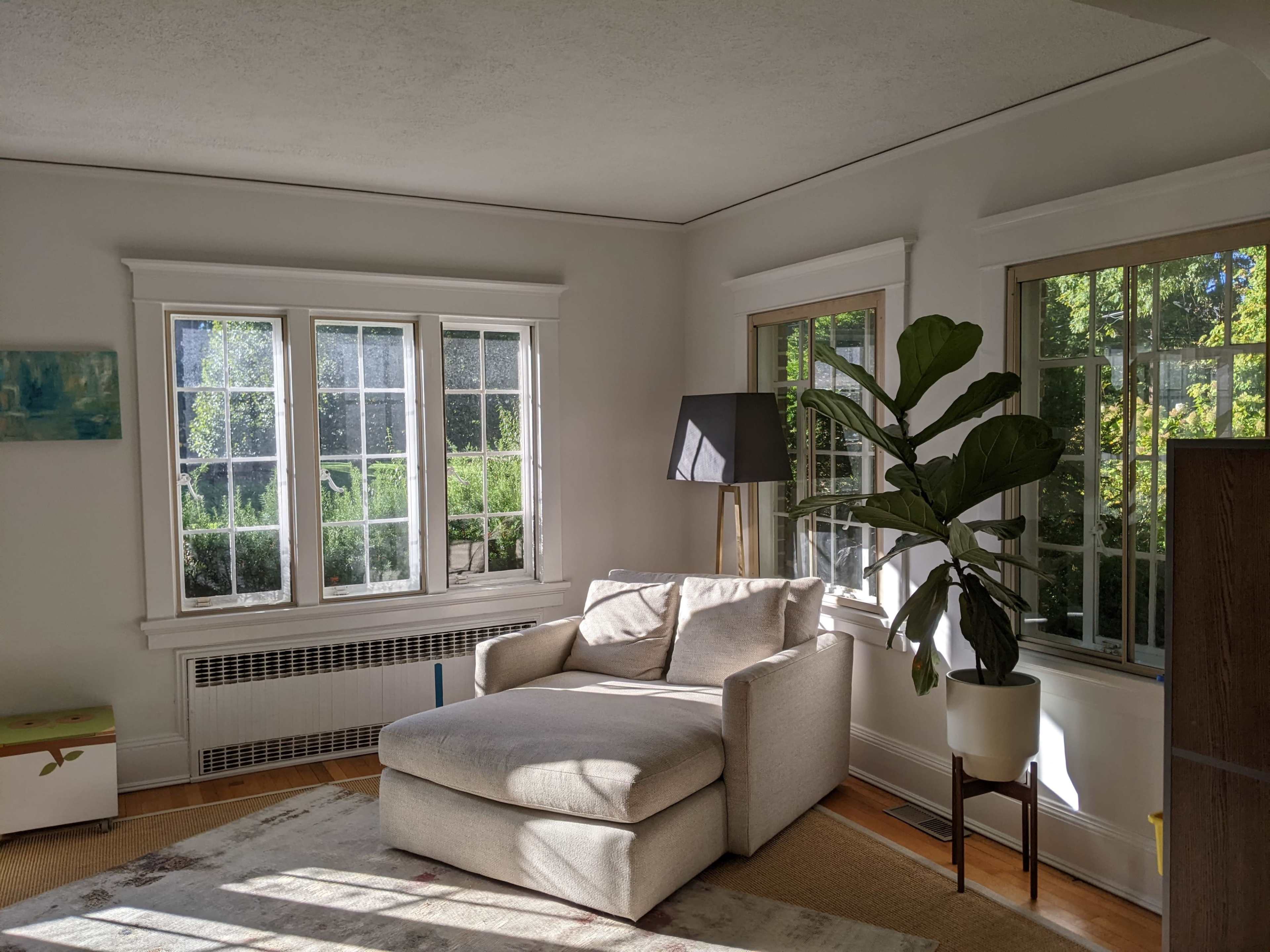 A well-lit living room with a light-colored armchair, a floor lamp, large windows, and a potted plant.