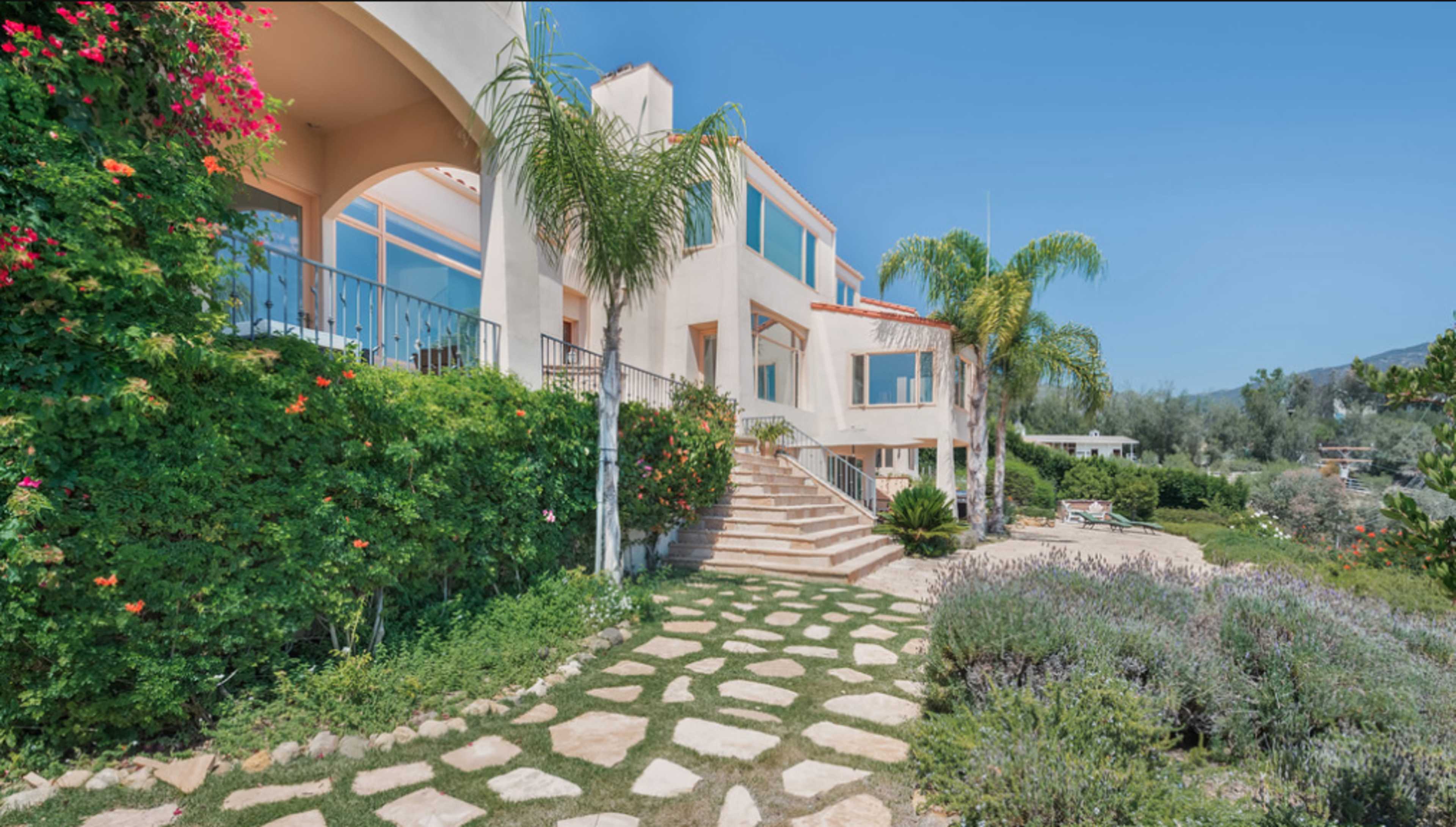 A modern house with a landscaped yard and stone pathway leads to a set of stairs flanked by palm trees.