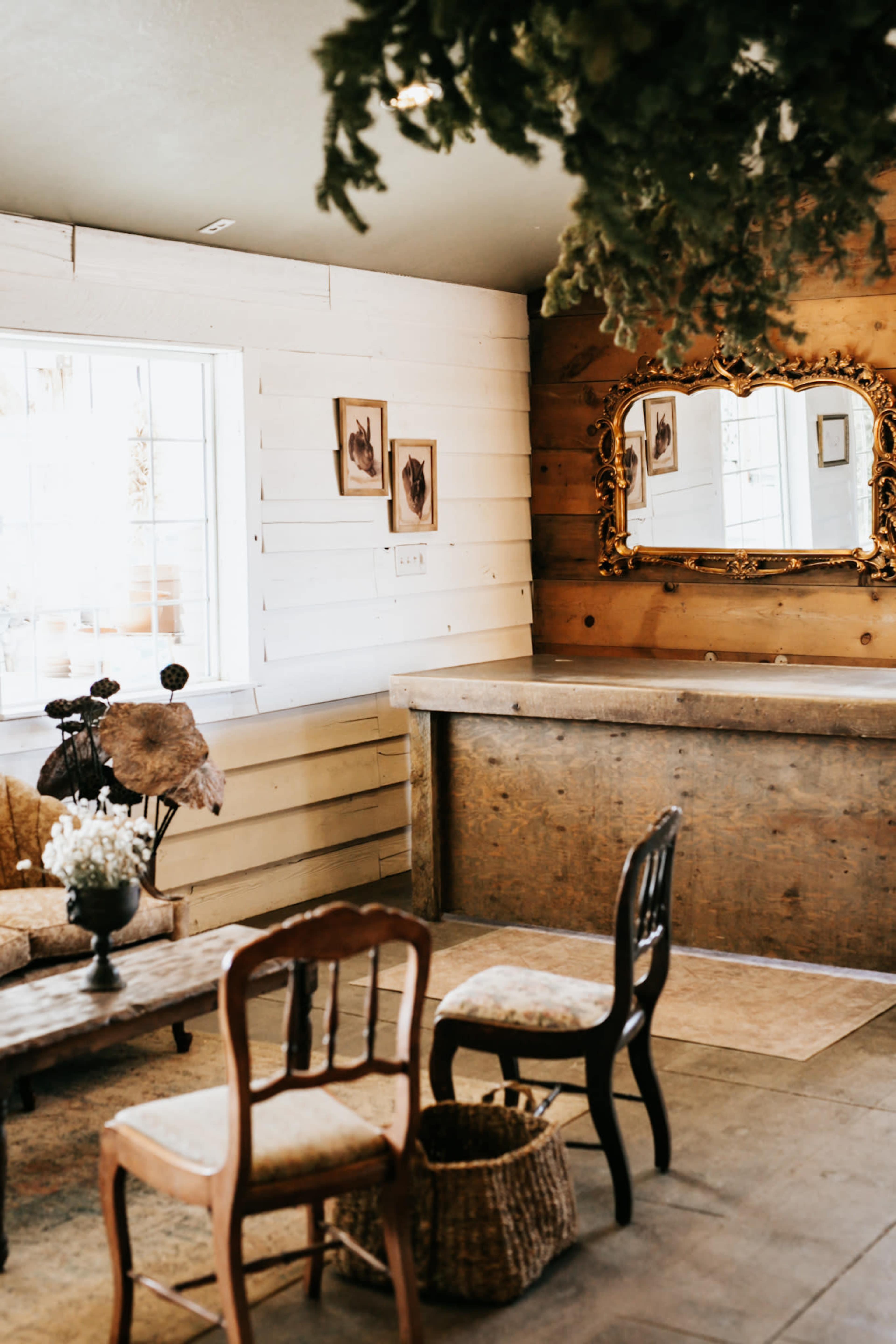 The image shows a welcoming interior featuring a wooden counter, several chairs, a rustic table, and framed pictures on the walls, with natural light coming through a window.