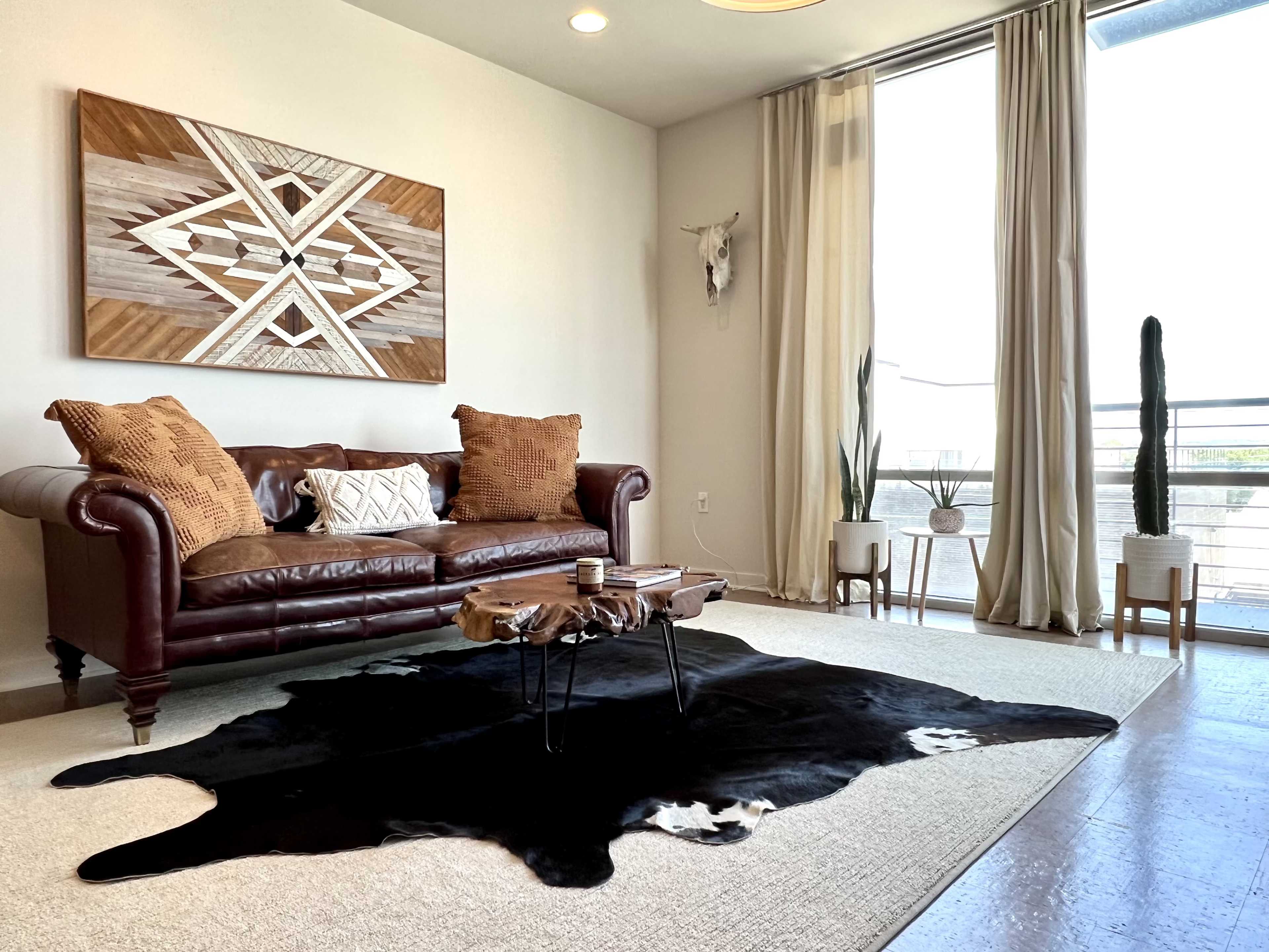 A modern living room featuring a brown leather sofa, a cowhide rug, wooden decor on the wall, and plants in decorative pots beside large windows.