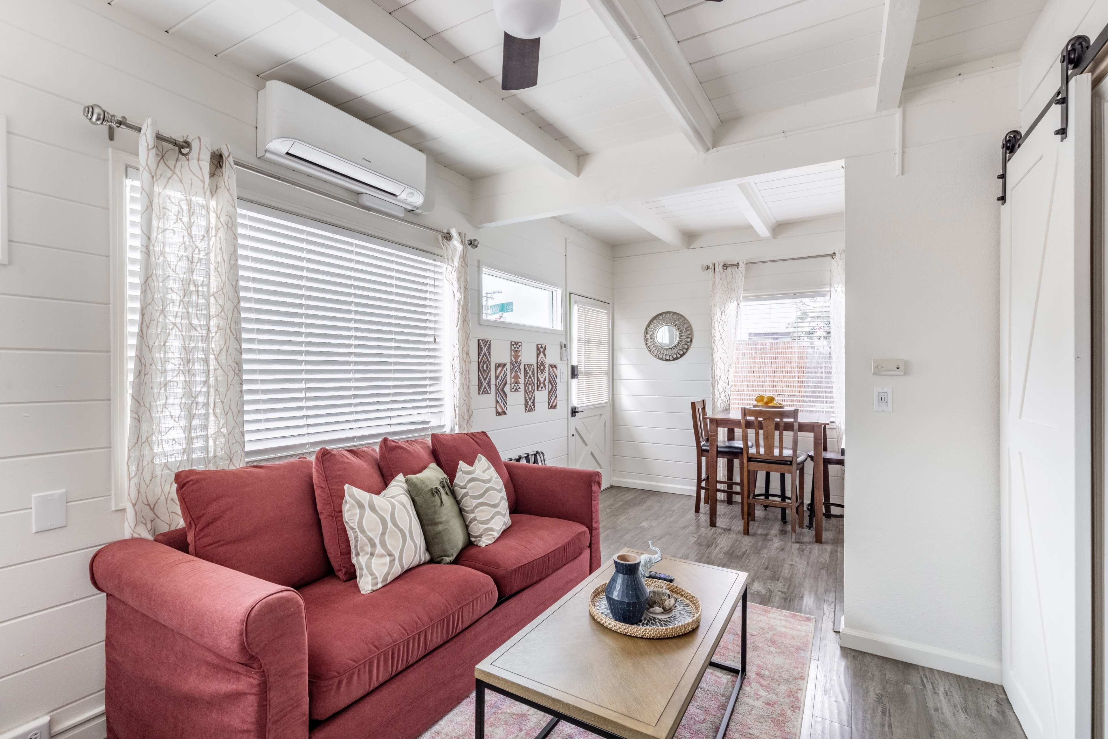 A bright, white interior of a living space with a red sofa, a coffee table, and a dining area with a wooden table and chairs.