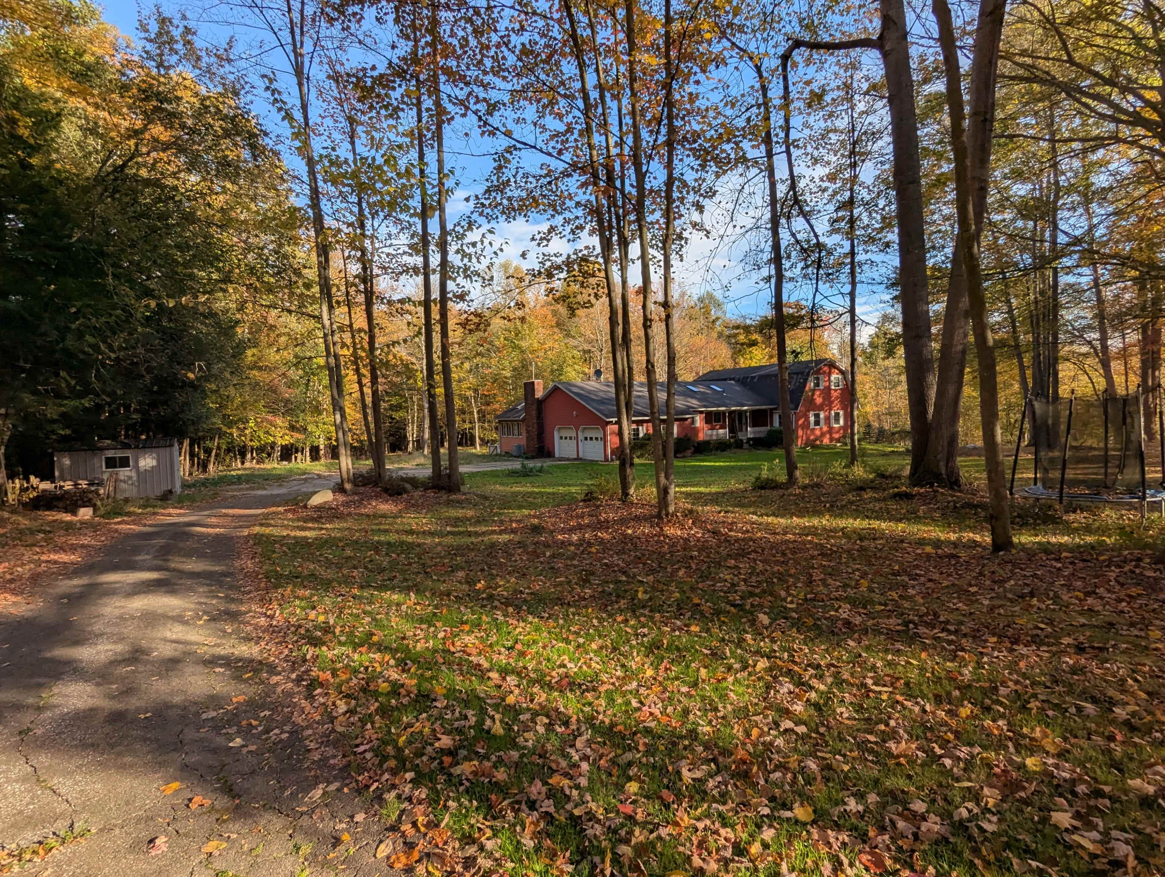 A gravel driveway leads to a red house surrounded by tall trees and autumn foliage.