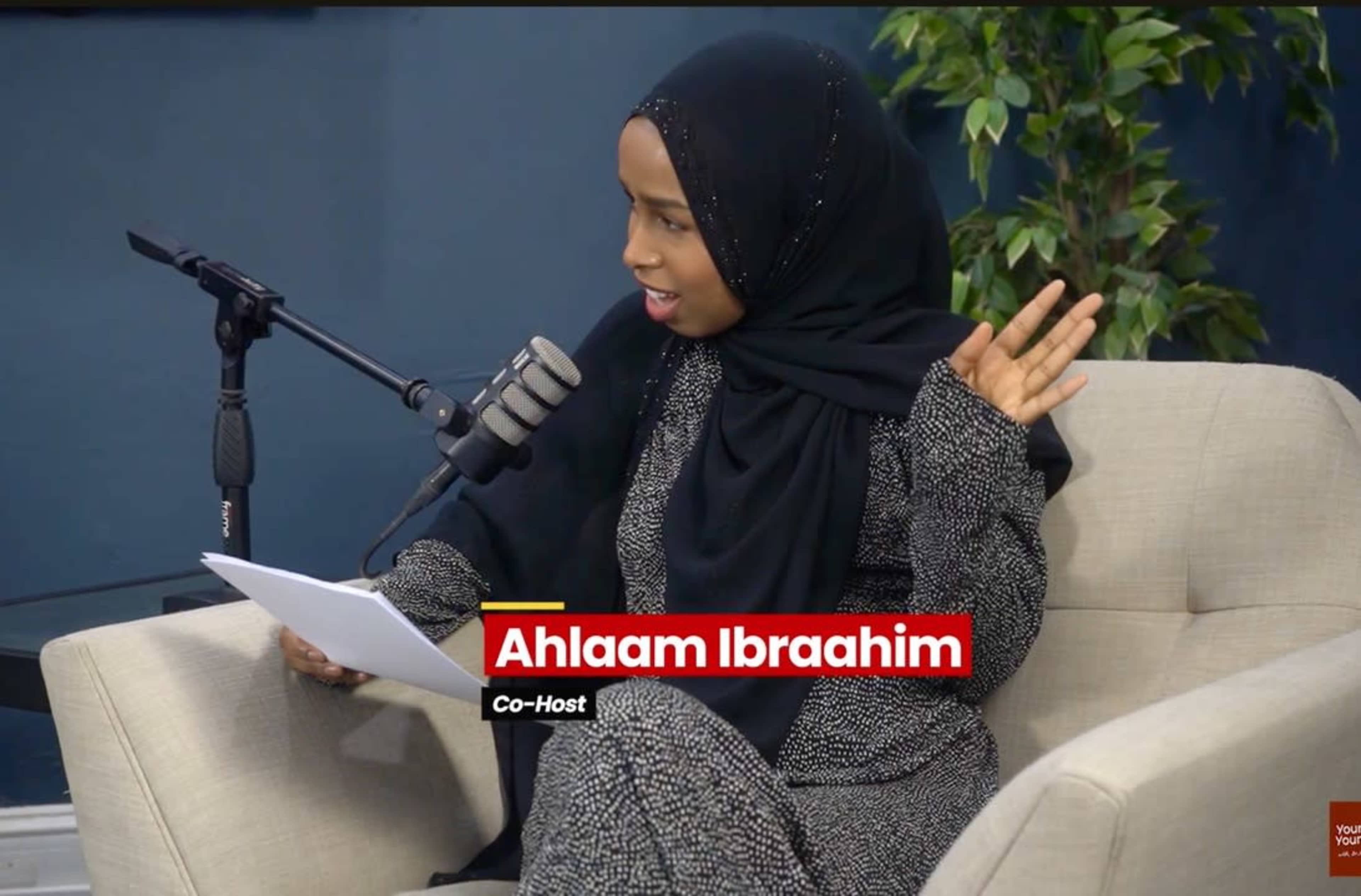 A woman wearing a hijab sits on a couch holding papers and speaks into a microphone during a recording session.