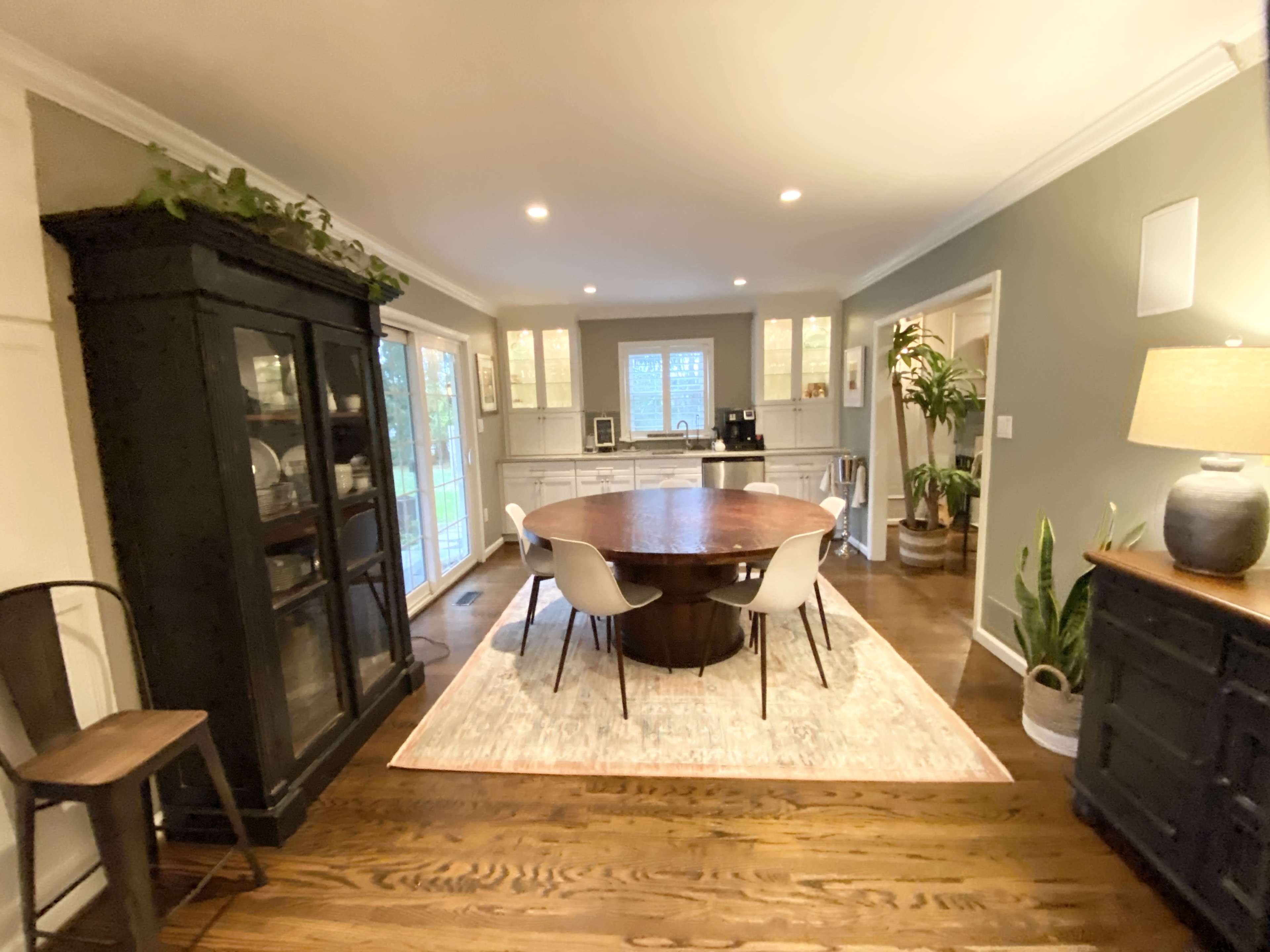 A modern dining area with a round wooden table surrounded by white chairs, a dark cabinet, and large windows letting in natural light.
