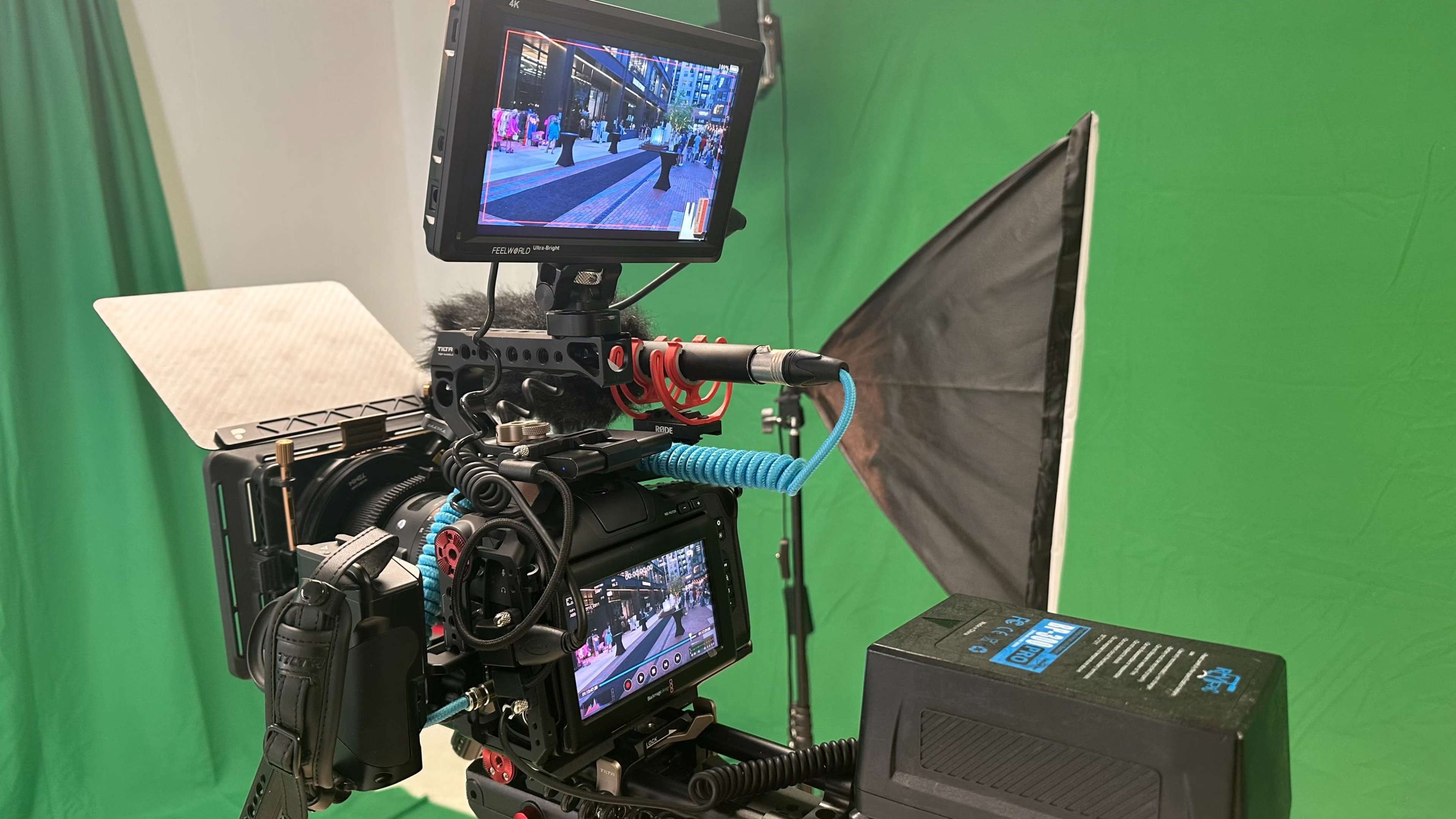 A video camera is set up in front of a green screen, displaying a live feed on its monitors.