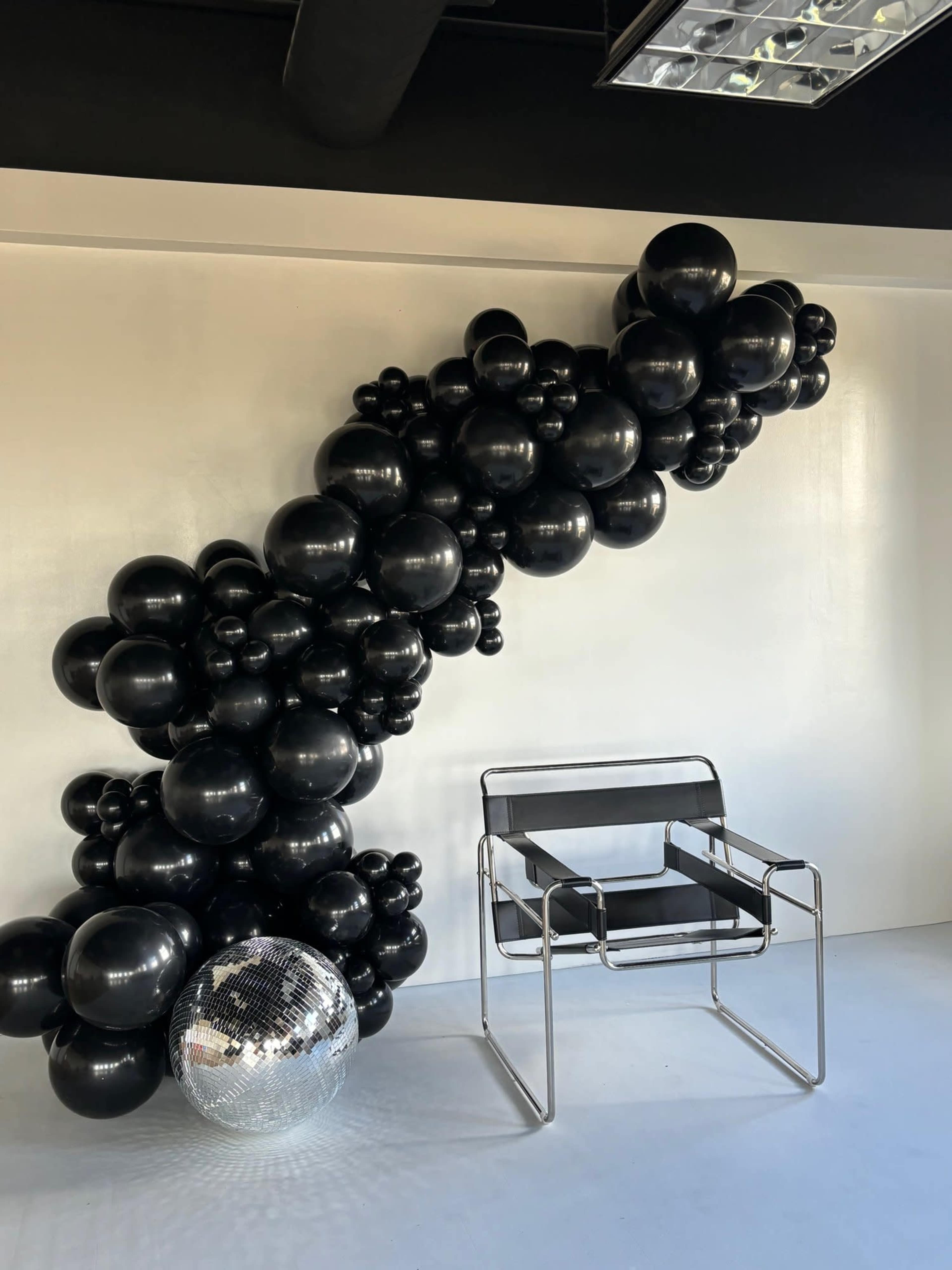 A cluster of black balloons forms a sculptural arrangement on a white wall, next to a modern metal chair and a disco ball on the floor.