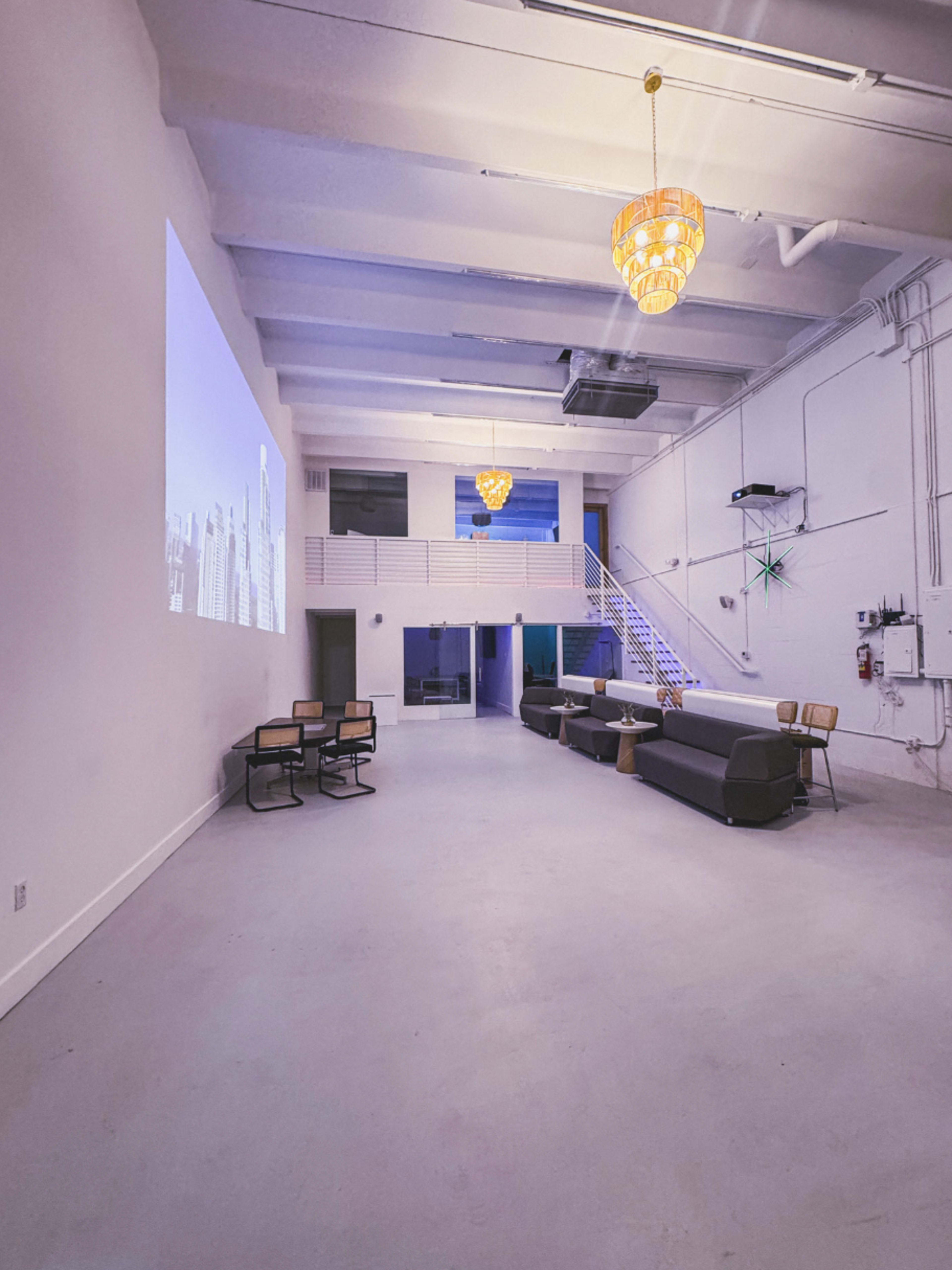 The image shows a spacious, modern interior with minimalist furniture, a projection screen on one wall, and decorative lighting fixtures hanging from the ceiling.