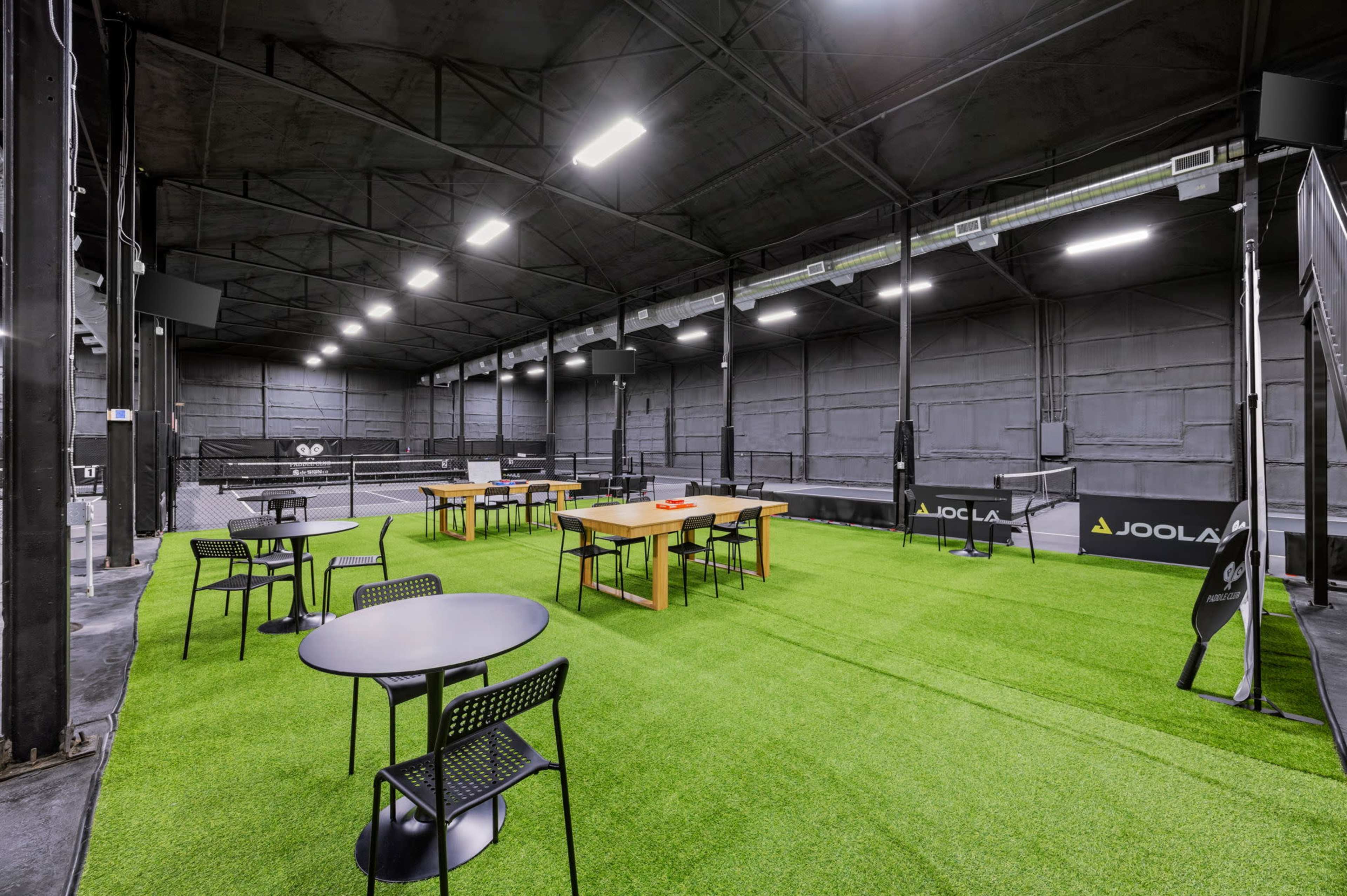 The image shows an indoor table tennis facility with several tables, surrounded by seating areas on green turf flooring.