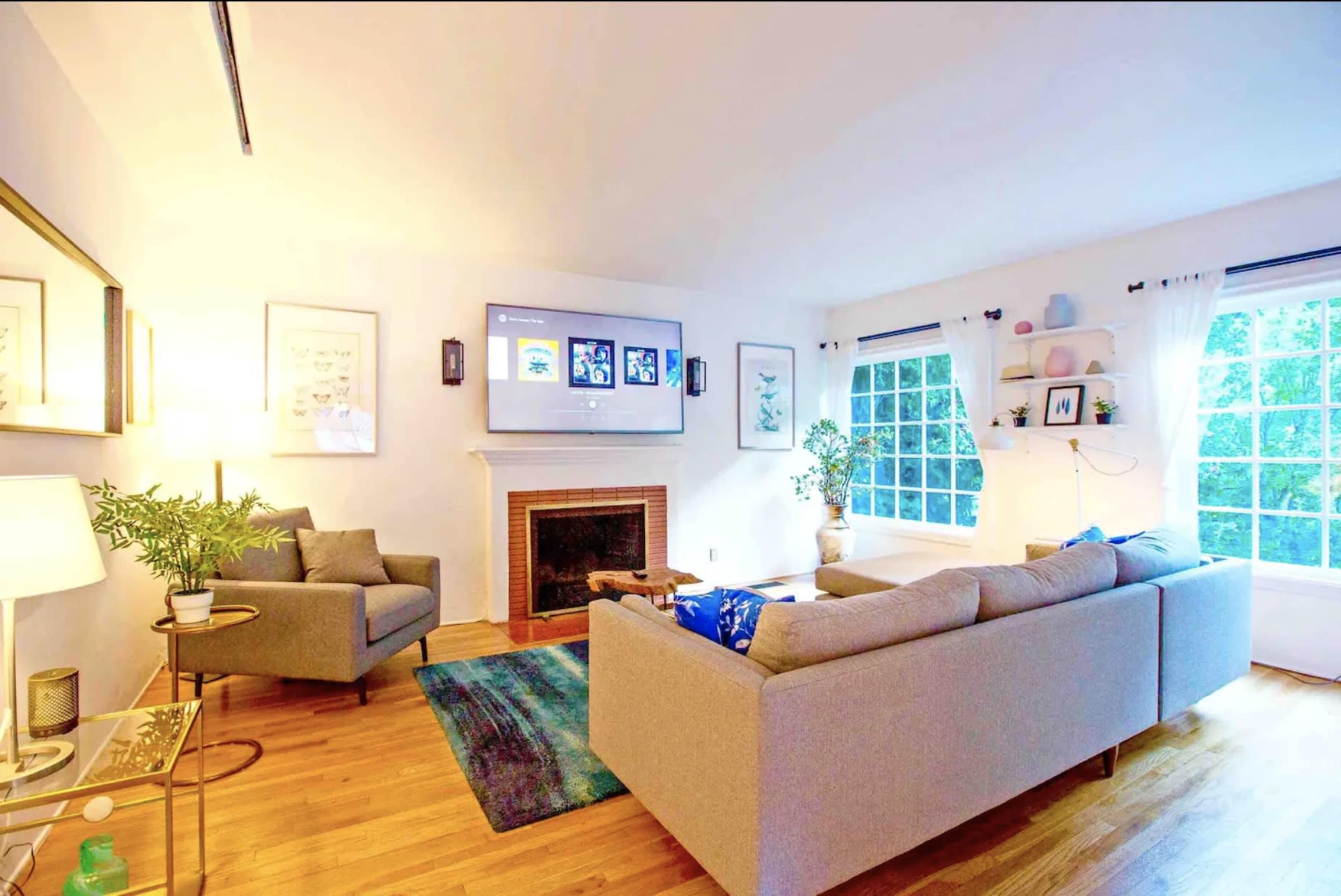 The image shows a modern living room with a gray sectional sofa, a fireplace, a TV mounted on the wall, and large windows with white curtains.