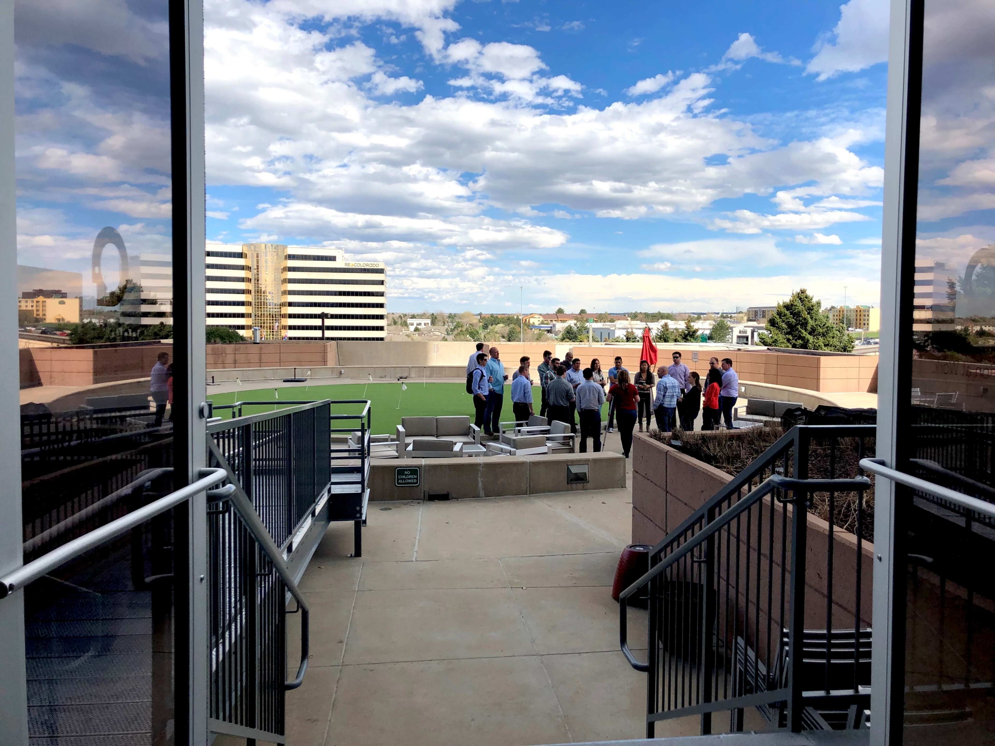 A group of people gathers on a rooftop space with greenery and buildings visible in the background under a partly cloudy sky.