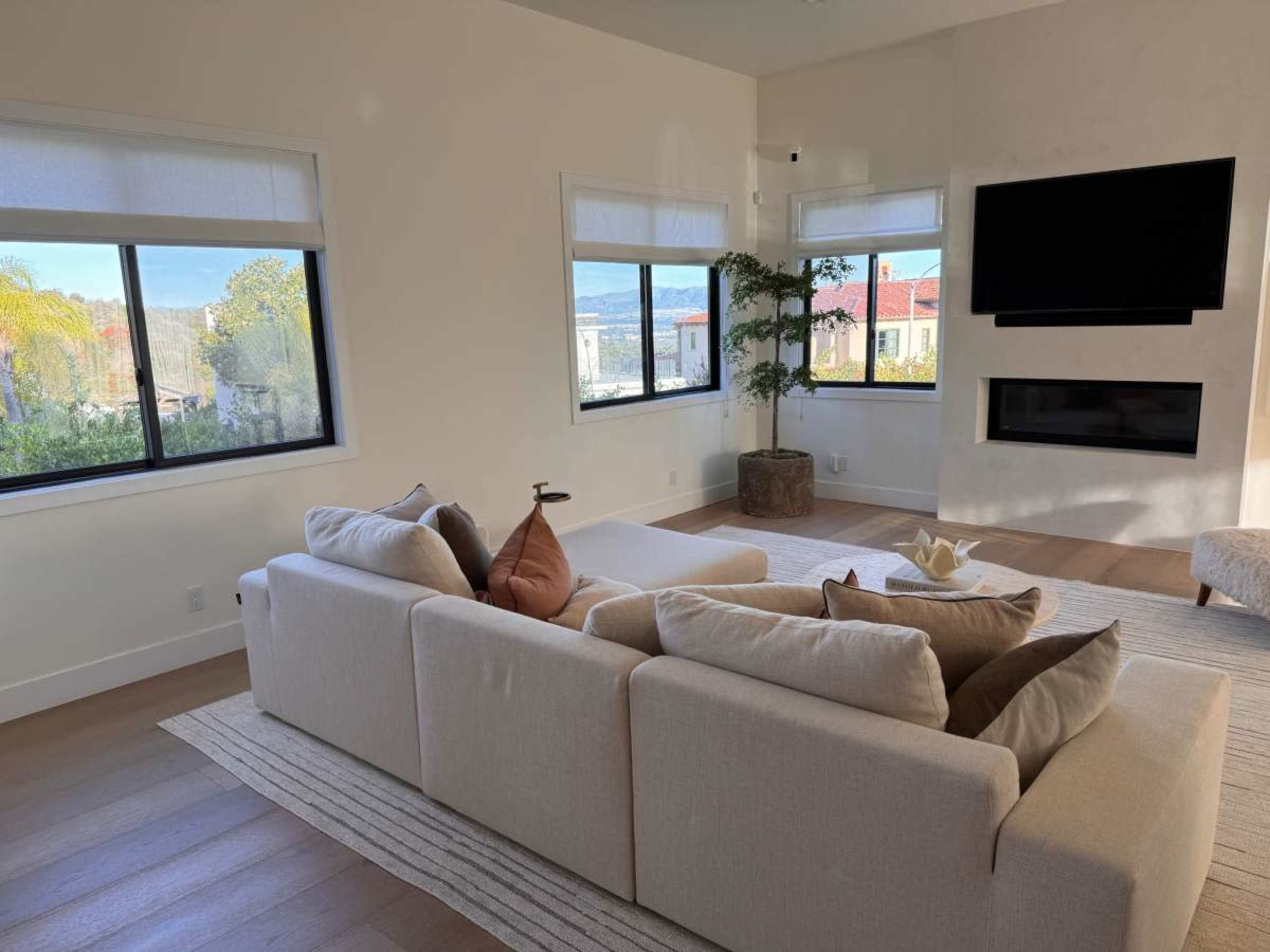 A modern living room features a light-colored sofa, a coffee table, large windows with views, and a mounted television on a neutral wall.