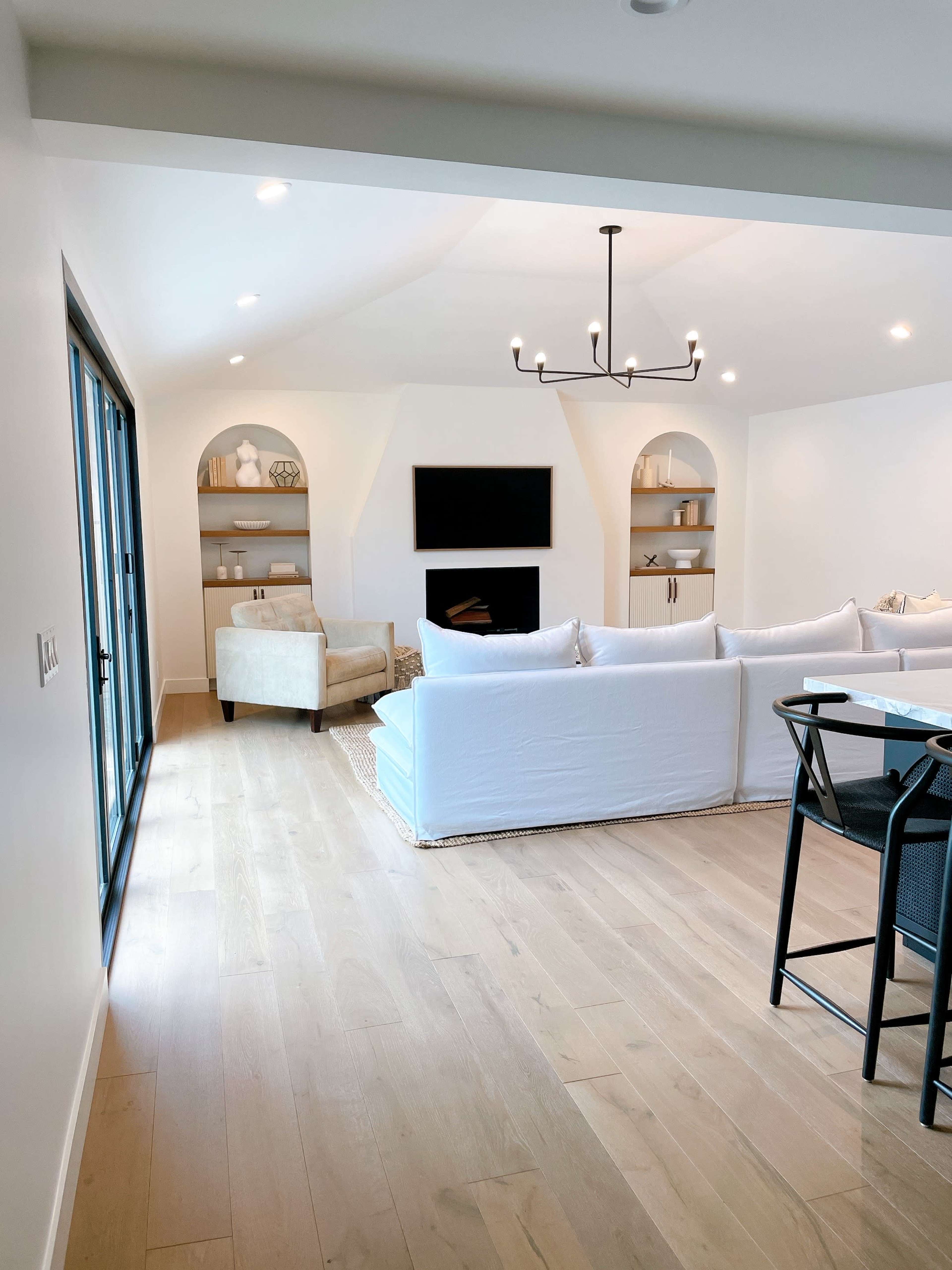 A bright, modern living space featuring a white sofa, a beige armchair, a wall-mounted television, and two built-in shelves with decorative items.