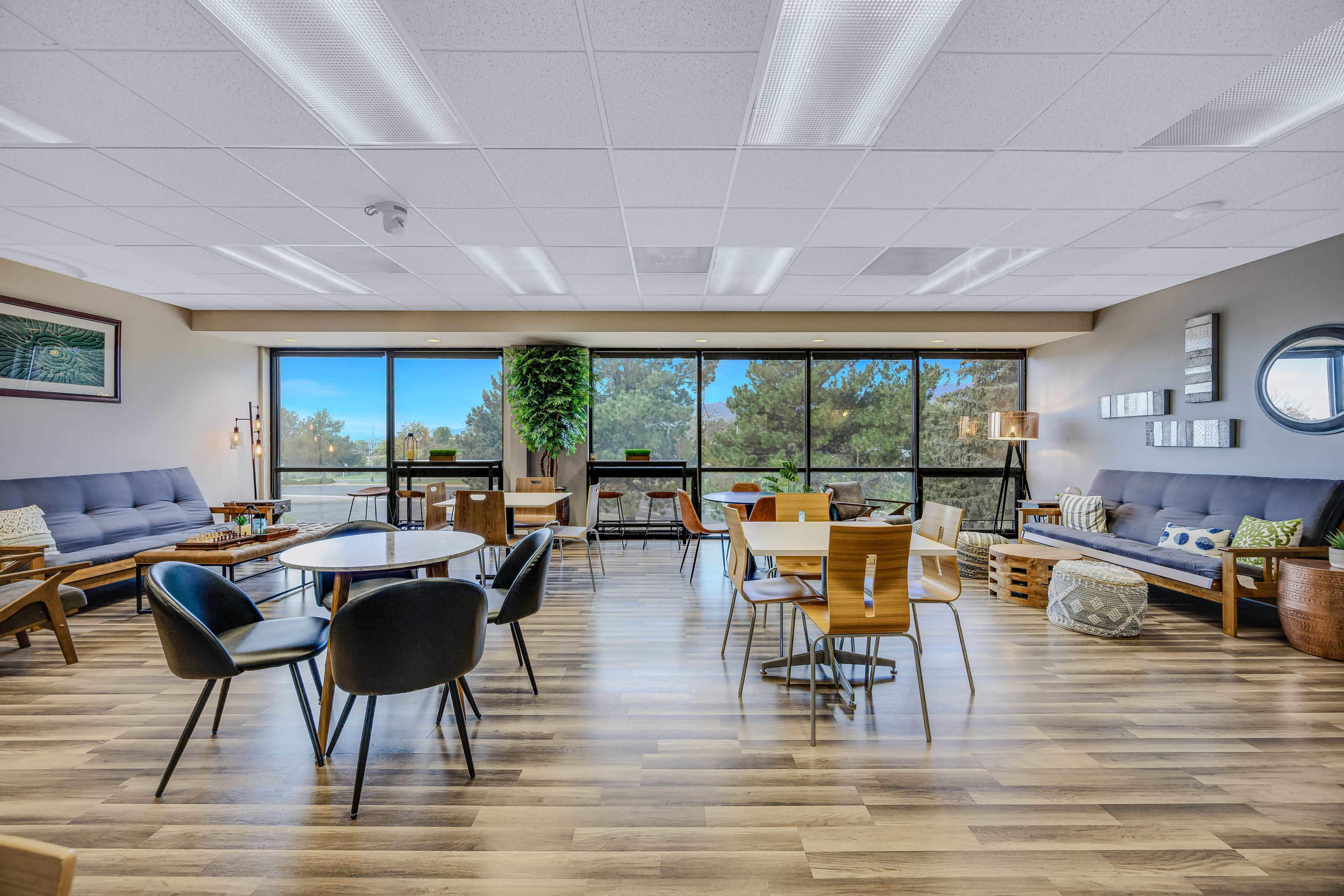 The image depicts a modern lounge area featuring seating arrangements with couches and chairs, a table, and large windows that offer a view of trees outside.