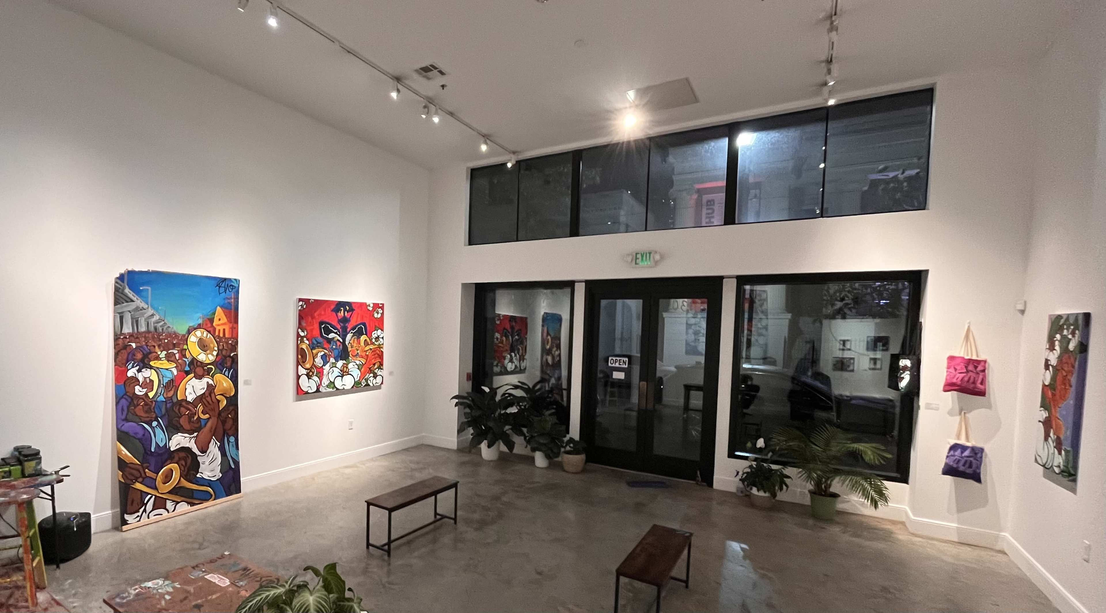 The image shows a modern art gallery featuring colorful paintings on the walls, a few small benches in the center, and large windows letting in natural light.