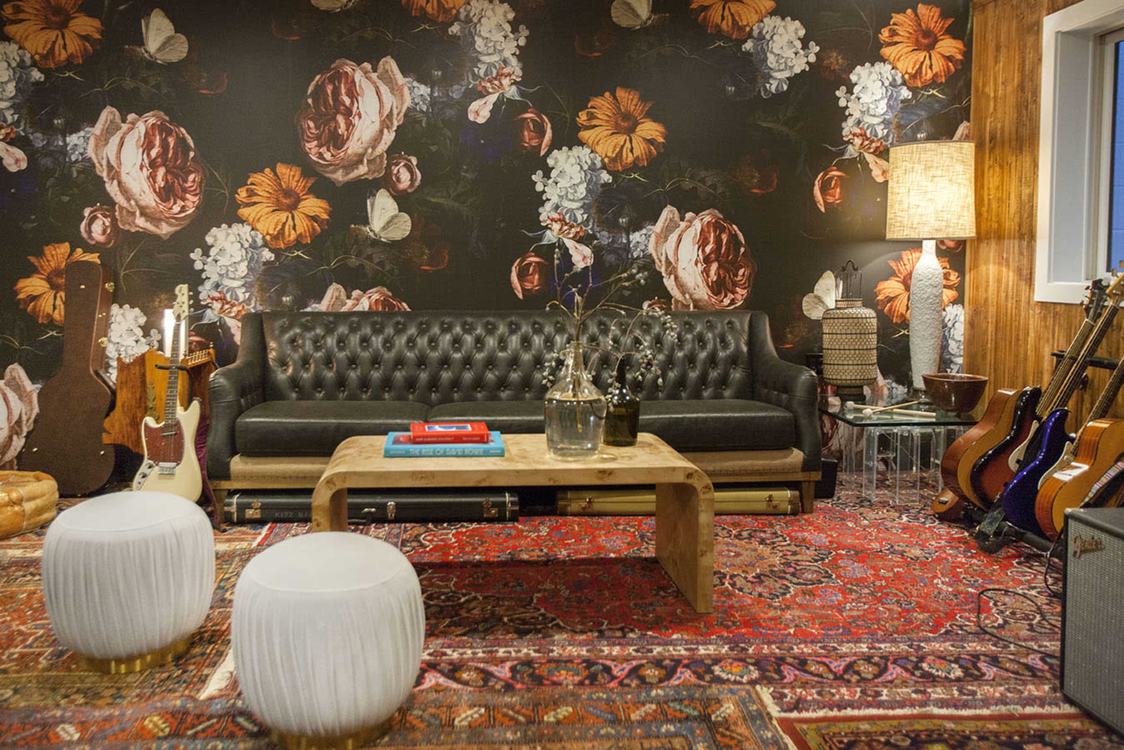 A living room featuring a black tufted leather sofa, a wooden coffee table, two white stools, a large floral wallpaper, and various musical instruments against the wall.