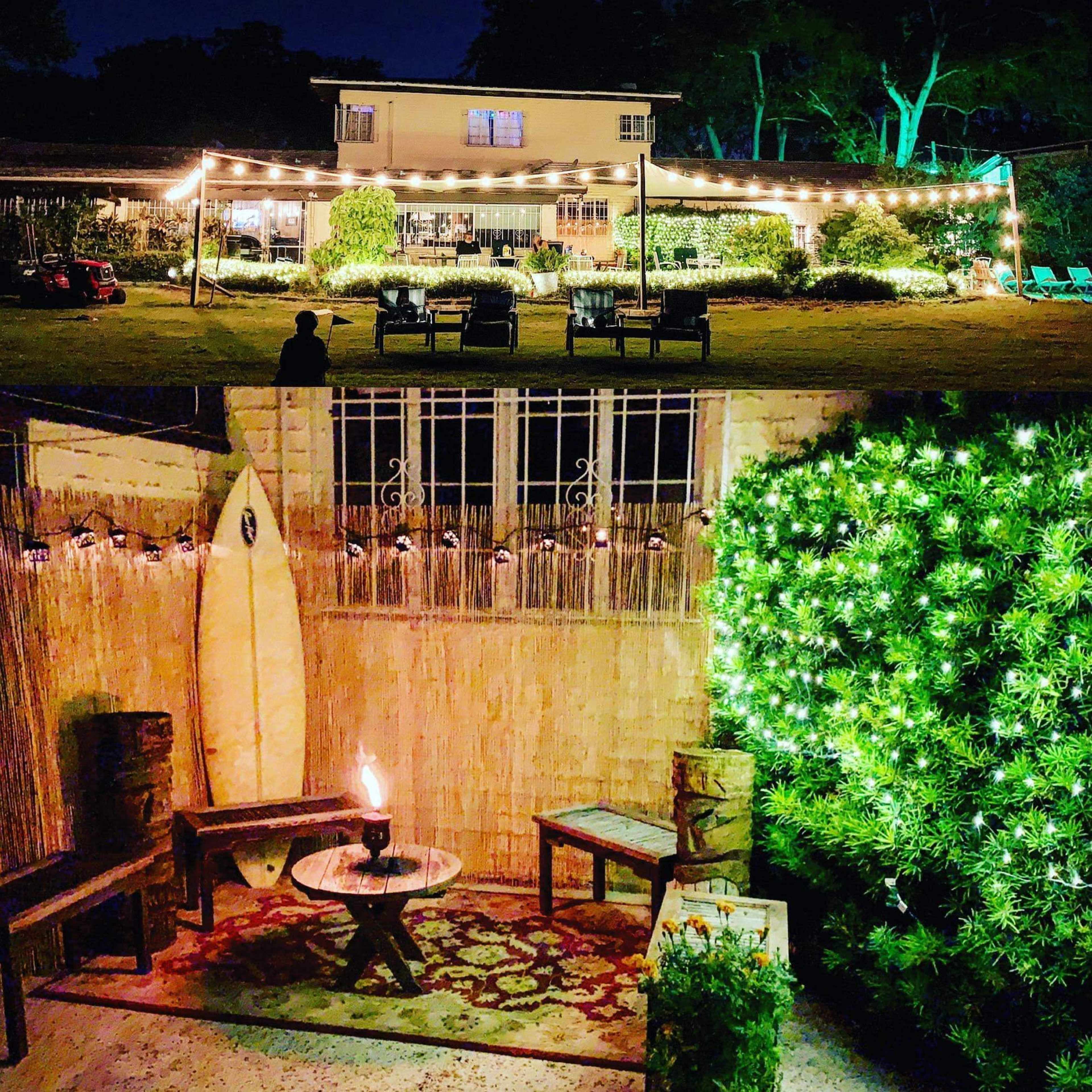 A backyard setting at night with string lights illuminating a seating area and a surfboard against a bamboo wall, surrounded by greenery.