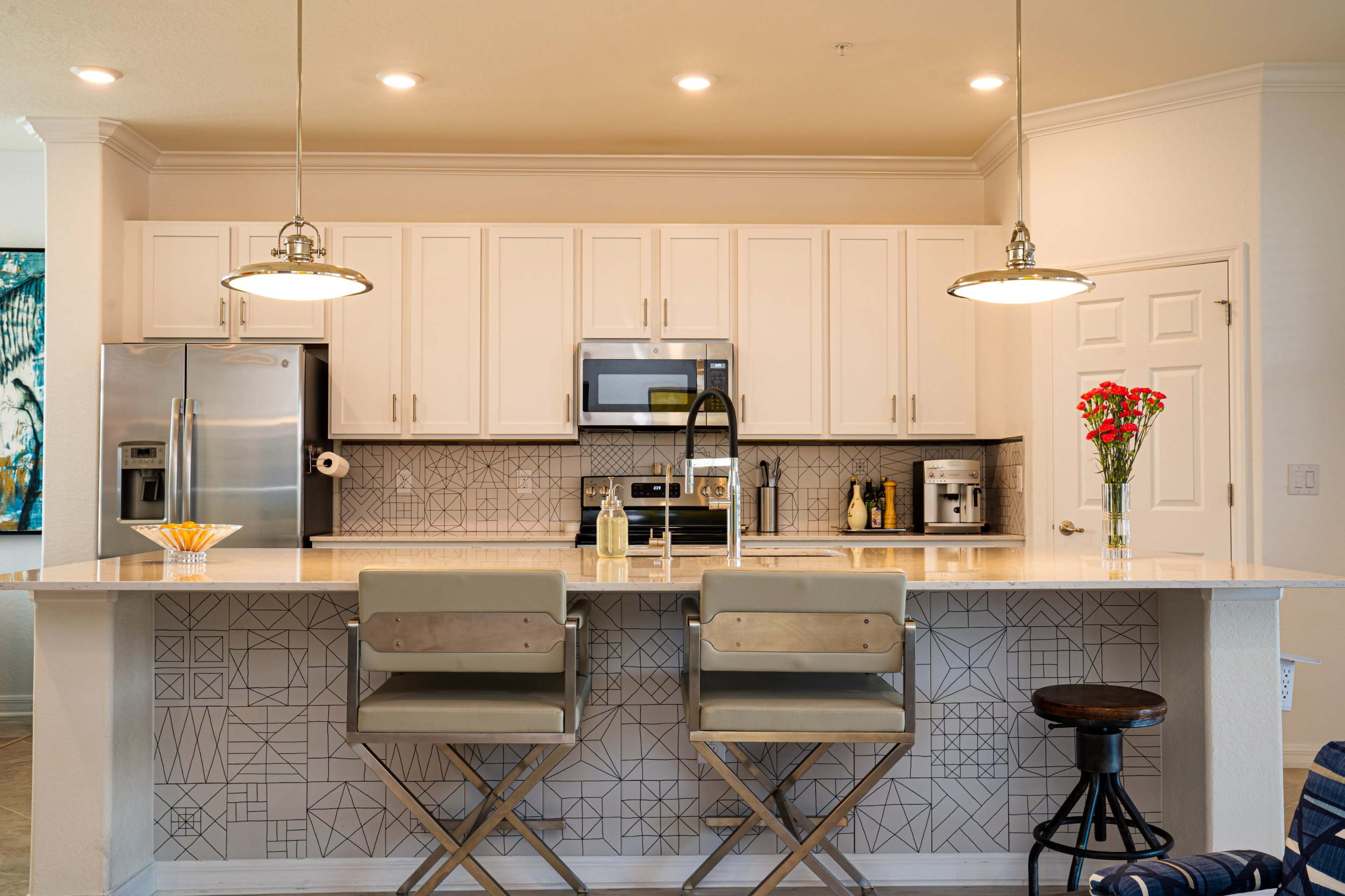 A modern kitchen with white cabinetry, stainless steel appliances, and a central island featuring bar stools and decorative tile work.