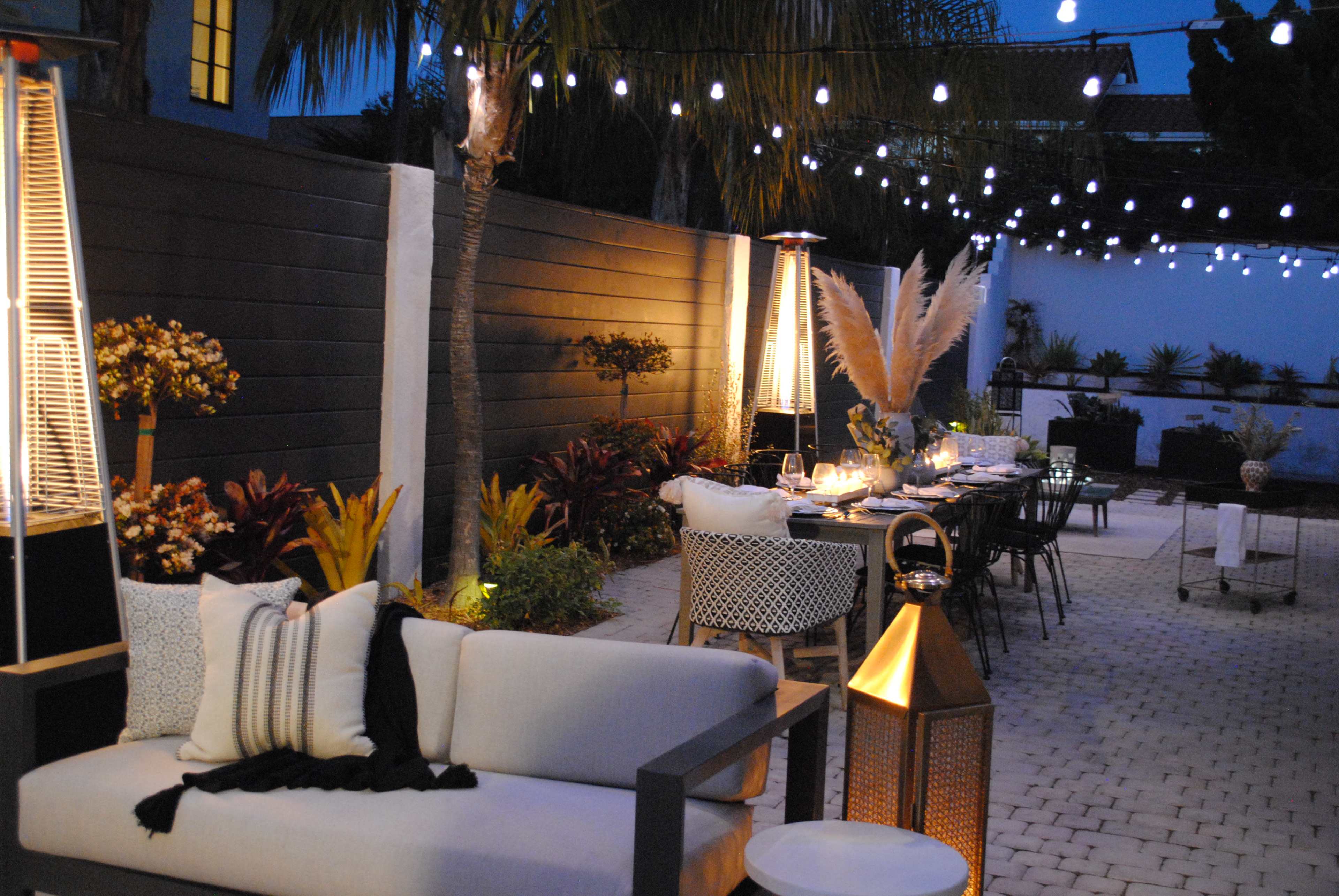 A modern outdoor dining area illuminated by string lights, featuring a long table set for dinner beside a lounge space with comfortable seating and decorative plants.