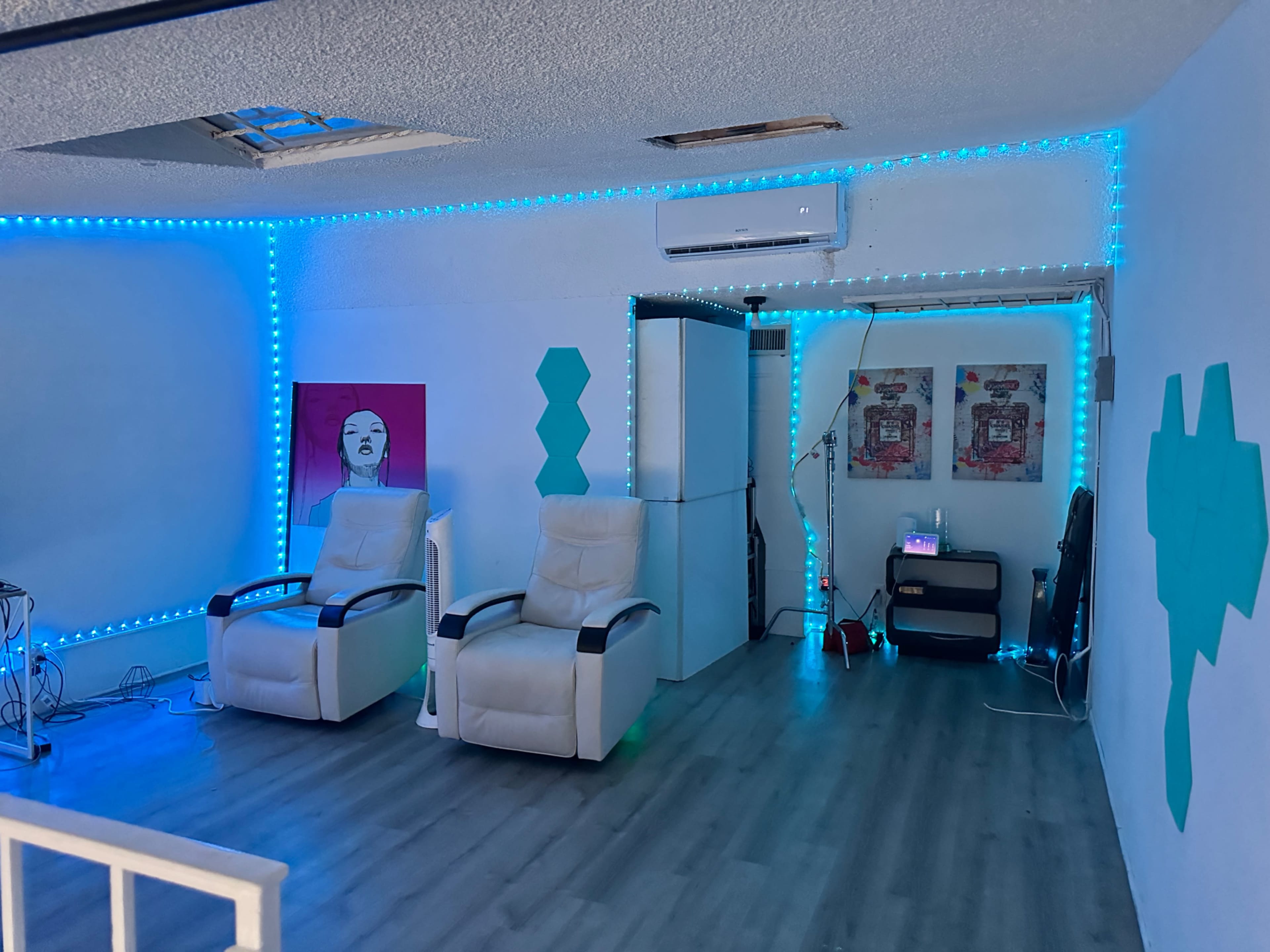 A brightly lit room with two white reclining chairs, blue LED strip lighting along the walls, and a mirrored area featuring colorful wall art.