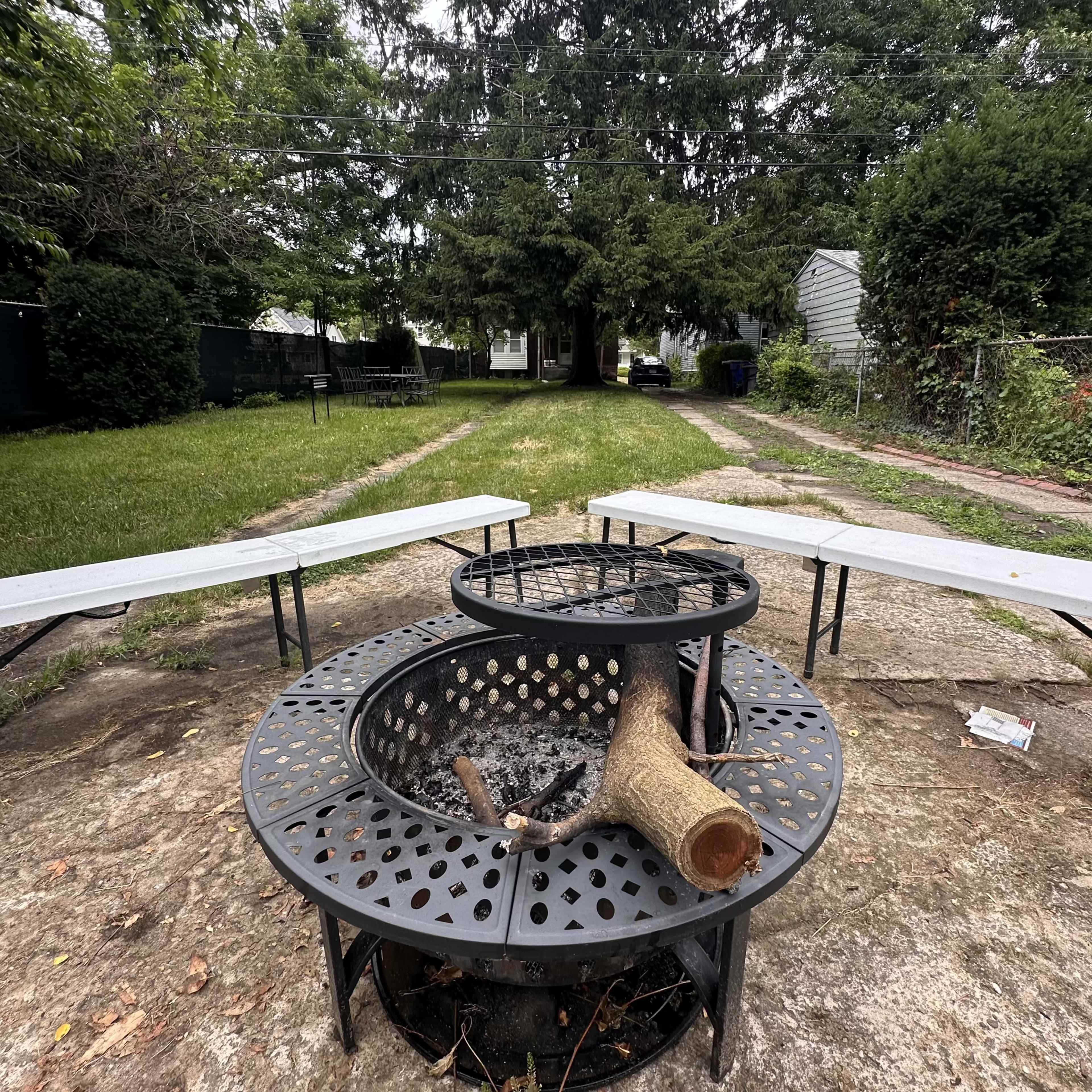 A metal fire pit with a log rests in the center of a grassy yard, surrounded by two folding tables arranged in a semi-circle.
