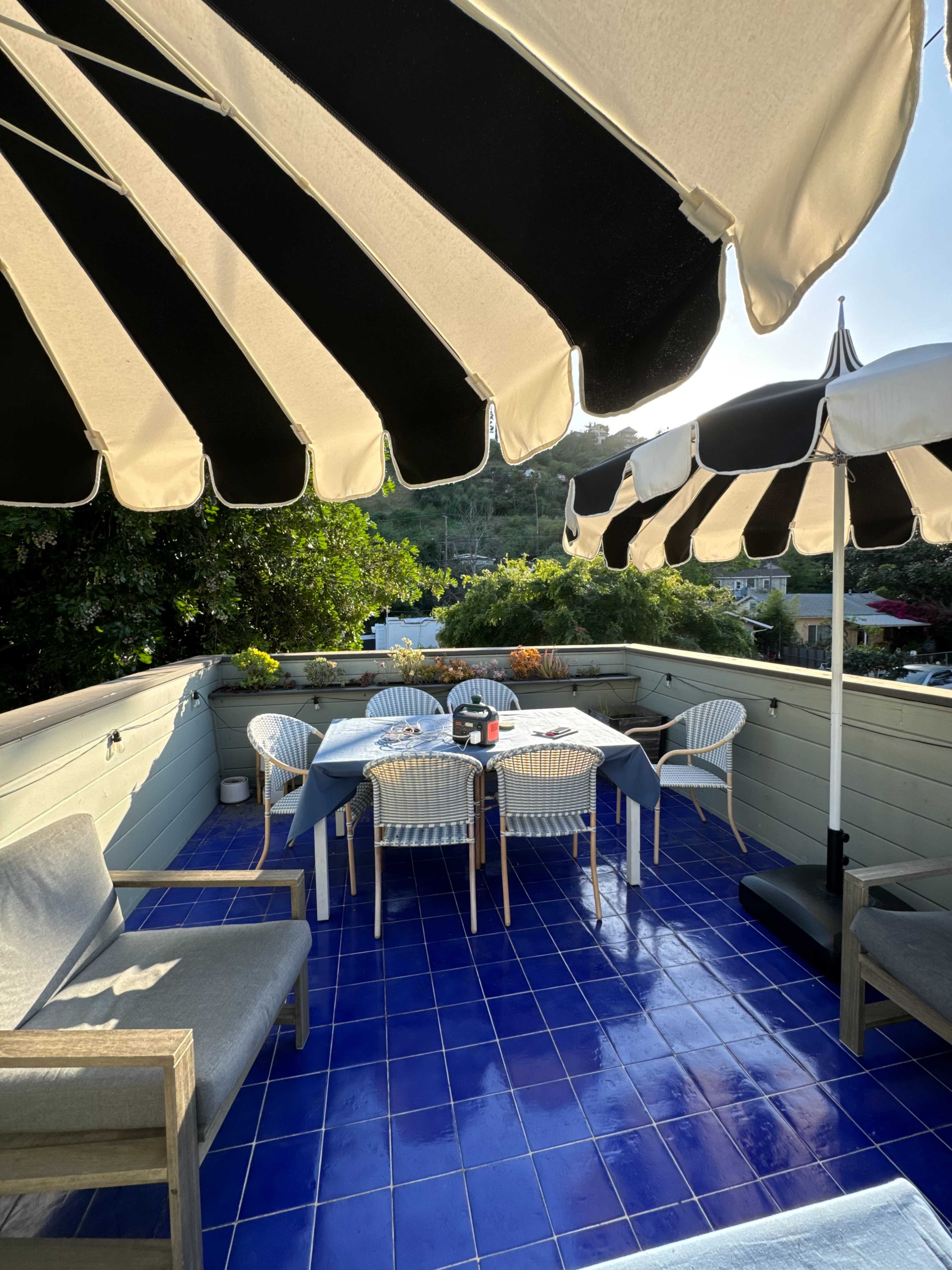 A rooftop terrace features a round dining table surrounded by striped umbrellas and blue tiled flooring.