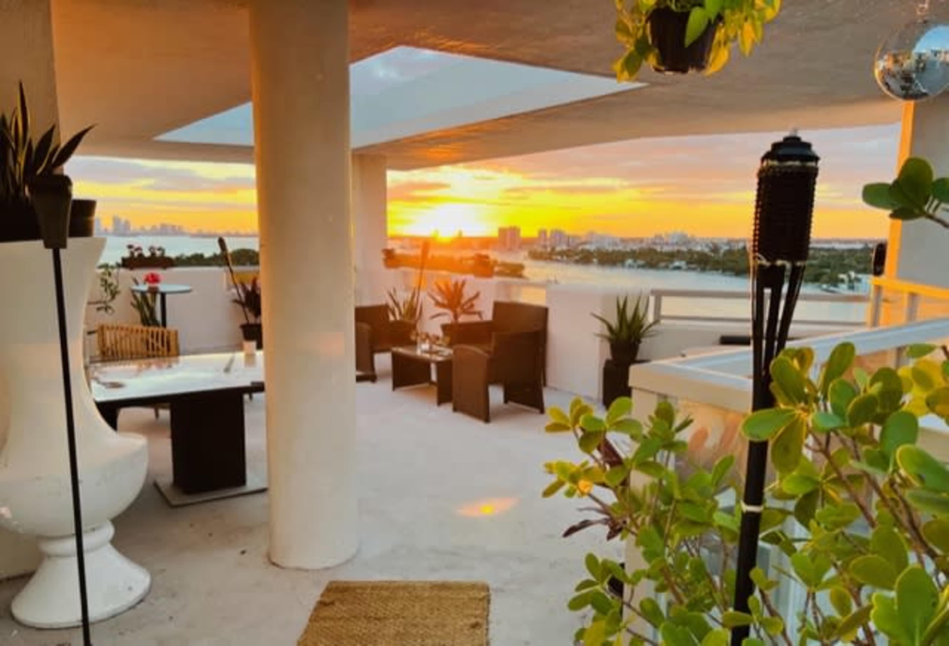 The image shows a rooftop terrace with seating arrangements and plants, overlooking a sunset in the background.