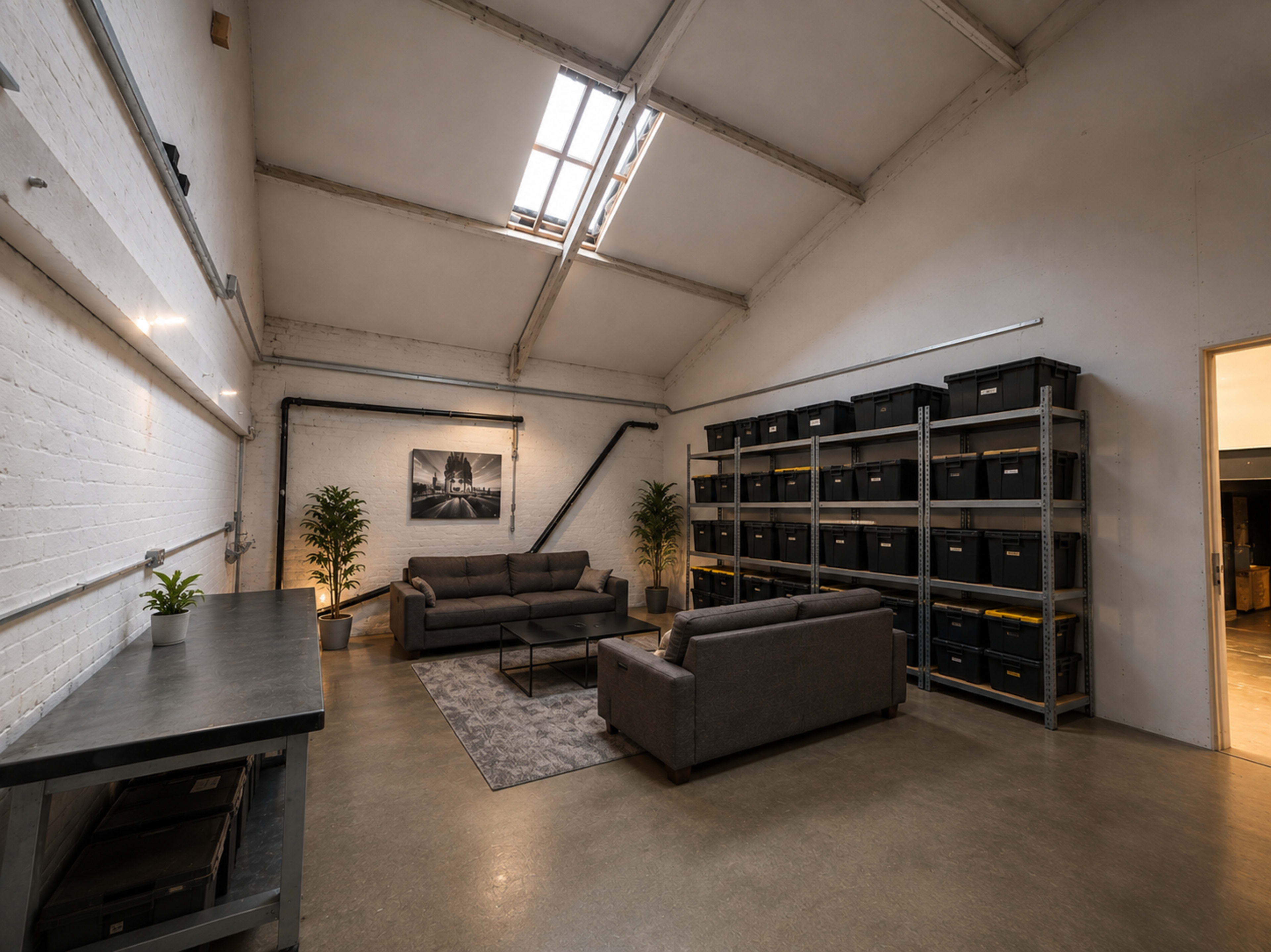 The image shows a spacious indoor area with a high ceiling, featuring two gray sofas, a coffee table, plants, and shelving units filled with black storage bins.
