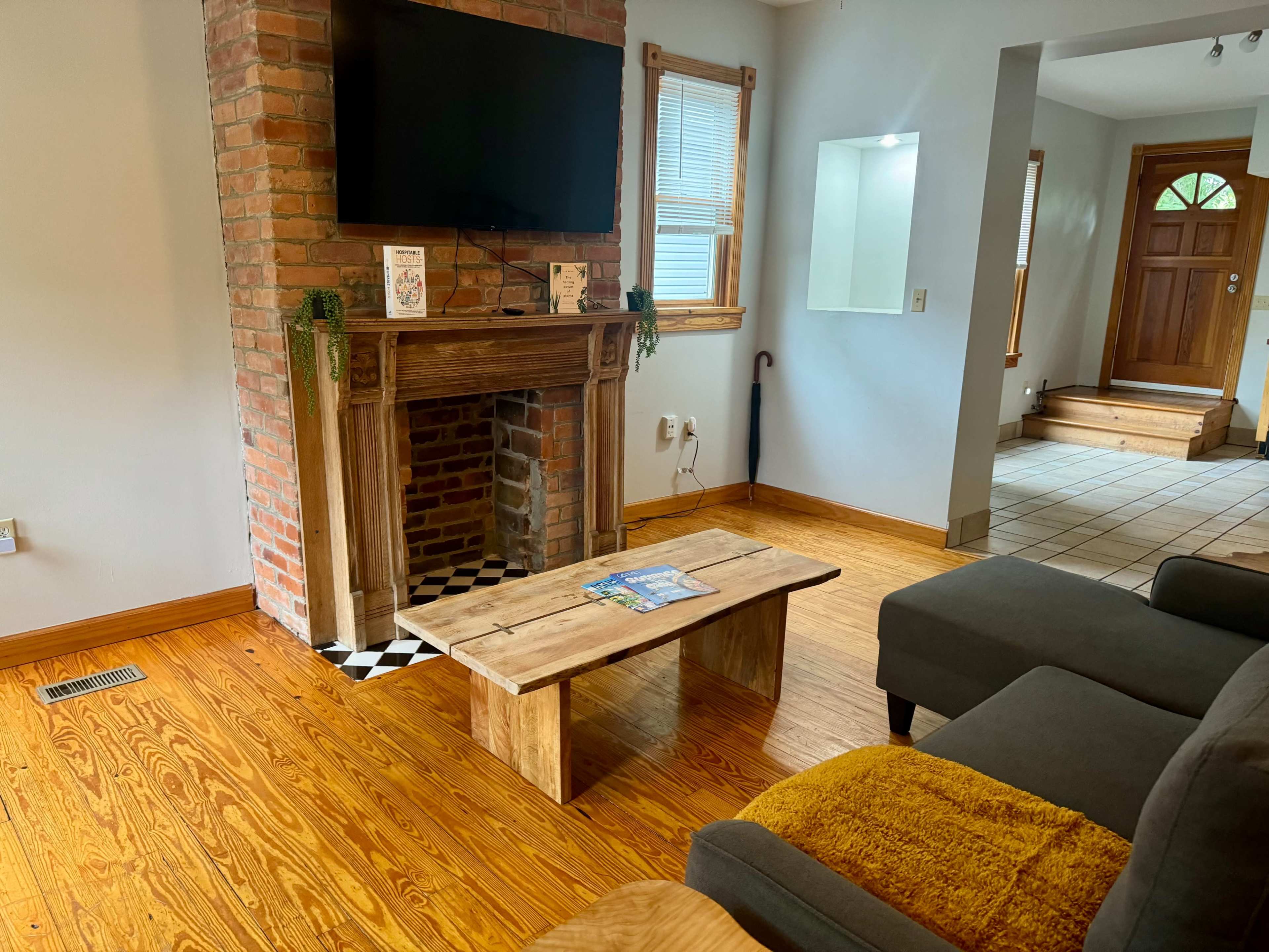 The living room features a brick fireplace with a television mounted above it, a wooden coffee table, and a gray couch, with a view of a door leading to an entrance.