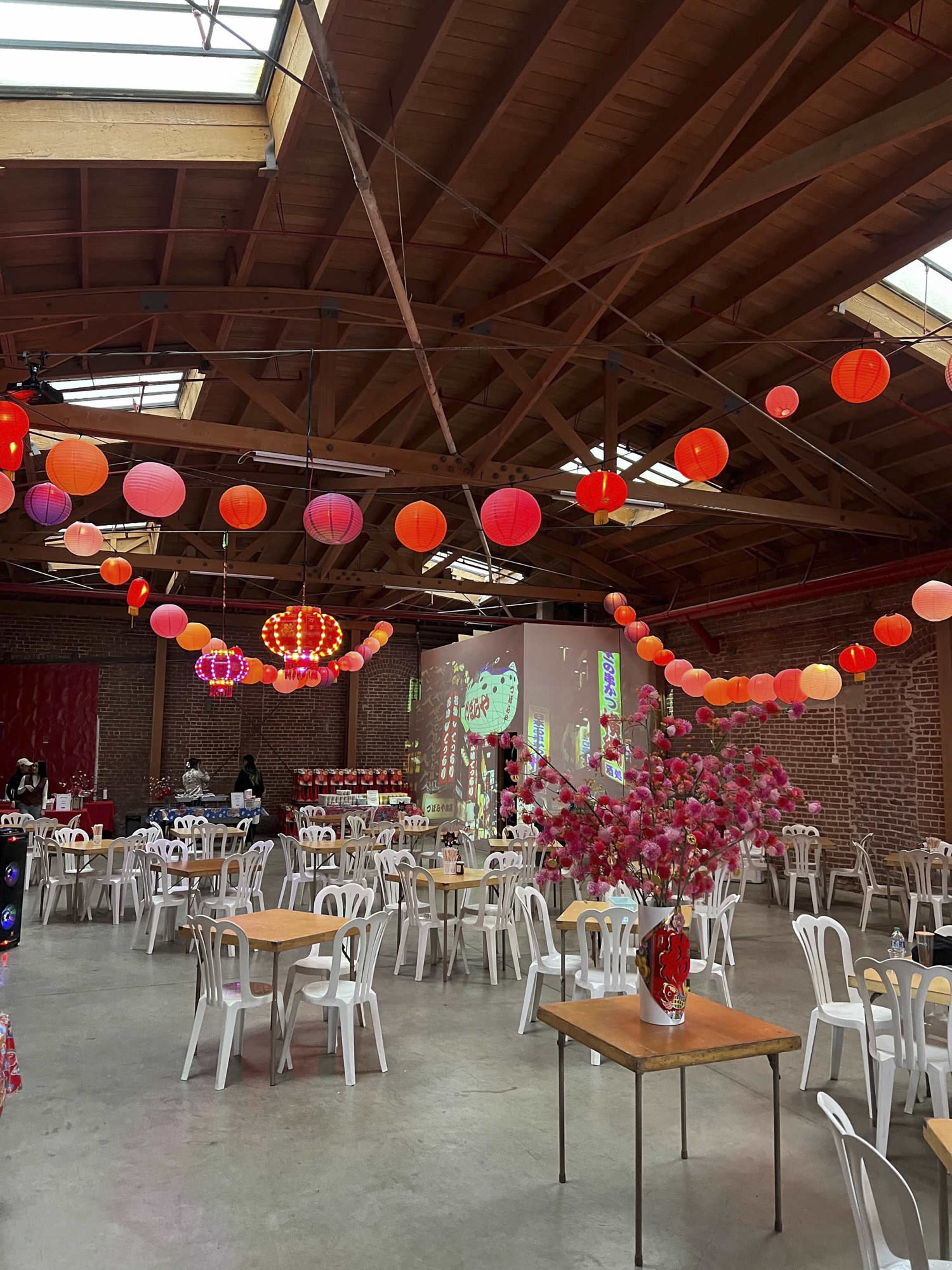 The image shows a spacious venue with wooden beams, decorated with colorful hanging lanterns and set with tables and chairs arranged for an event.
