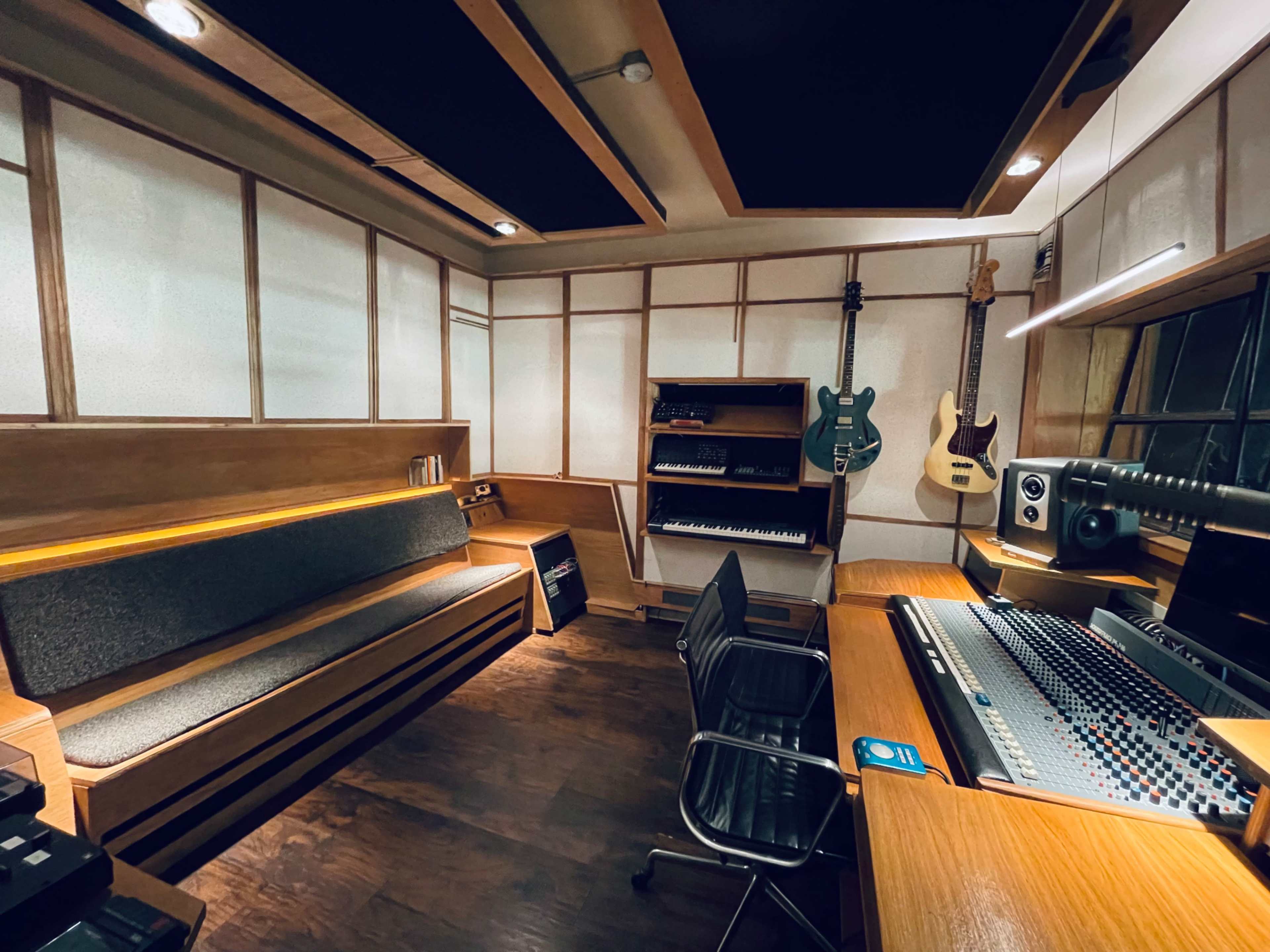 A music studio featuring a mixing console, monitors, and various instruments on the walls, with a wooden bench and shelving units.