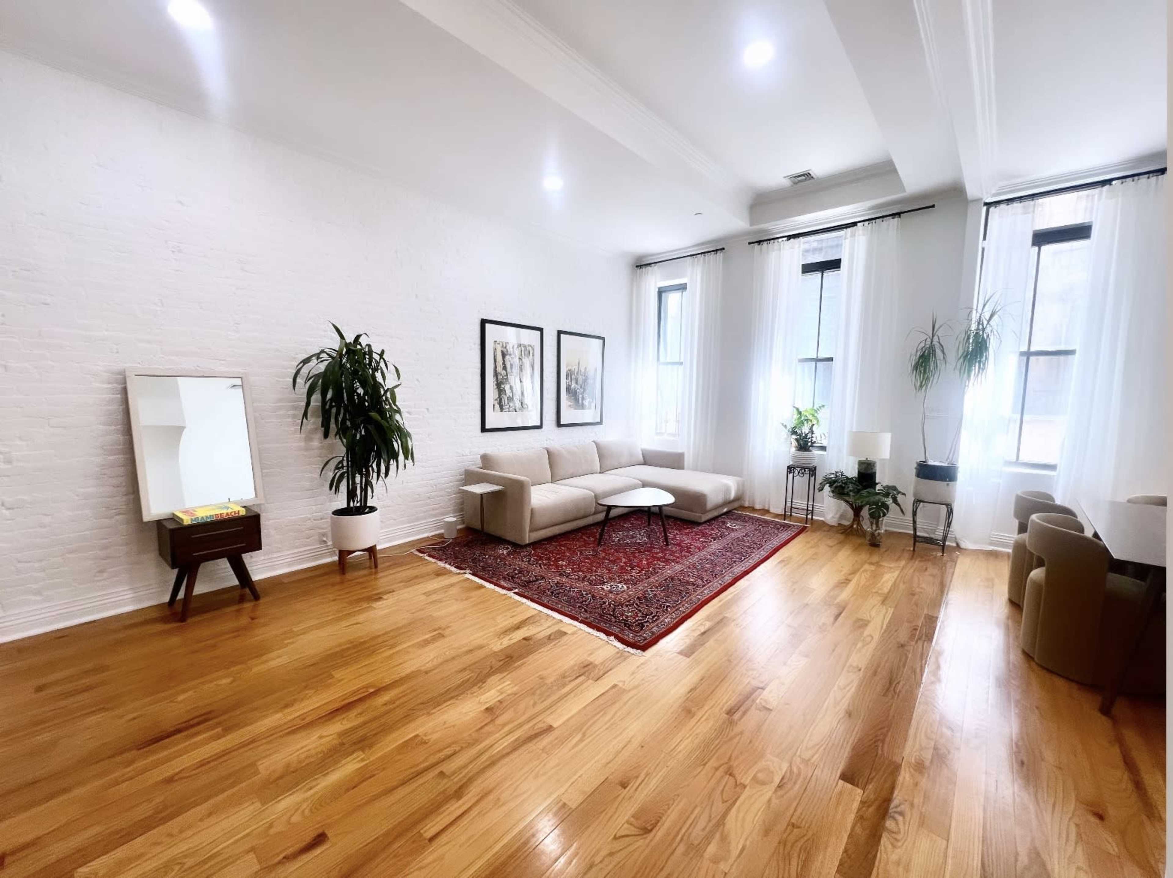 A spacious living room features a light-colored sectional sofa, a large area rug, framed artwork on the walls, and natural light streaming through tall windows.