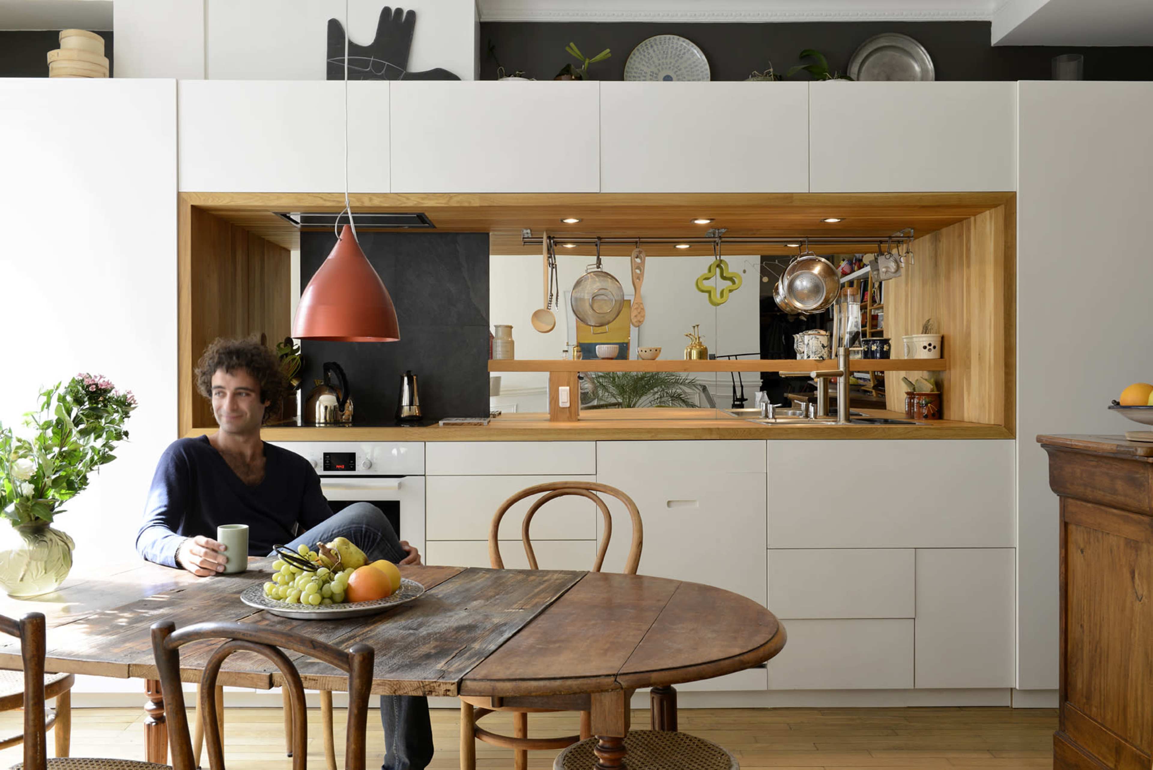 A man sits at a wooden dining table with a fruit bowl in front of him, while an open kitchen with a minimalist design and wooden accents is visible in the background.