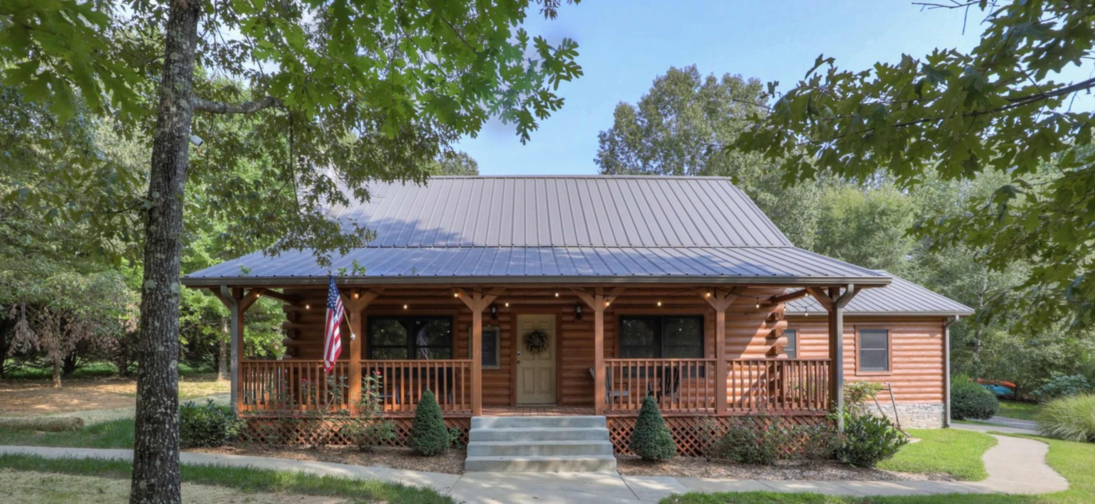 A log cabin-style house with a metal roof, surrounded by trees and featuring a front porch with decorative railing and a flag.