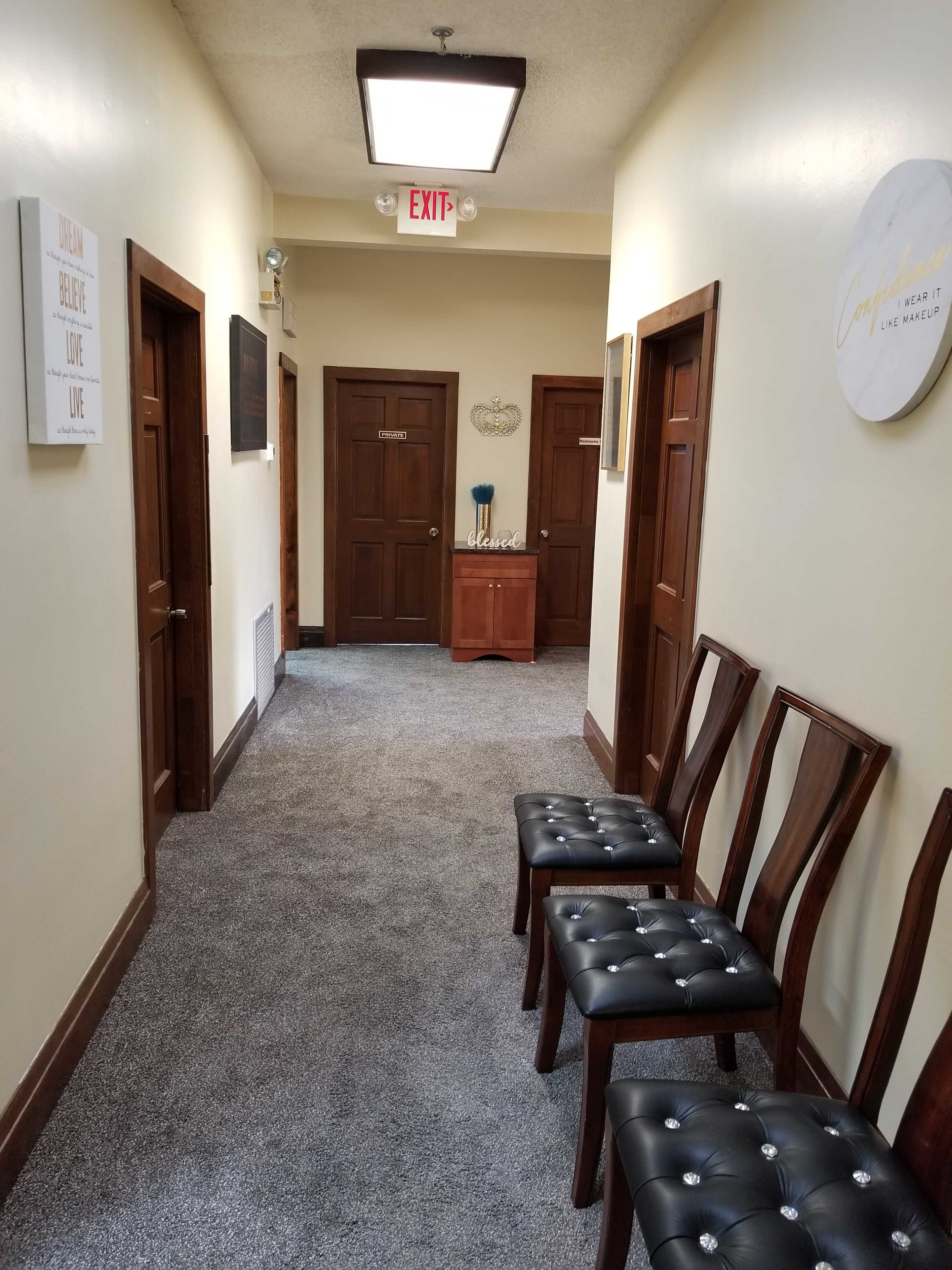 A hallway with light-colored walls, wooden doors on either side, and a row of four wooden chairs positioned against one wall.