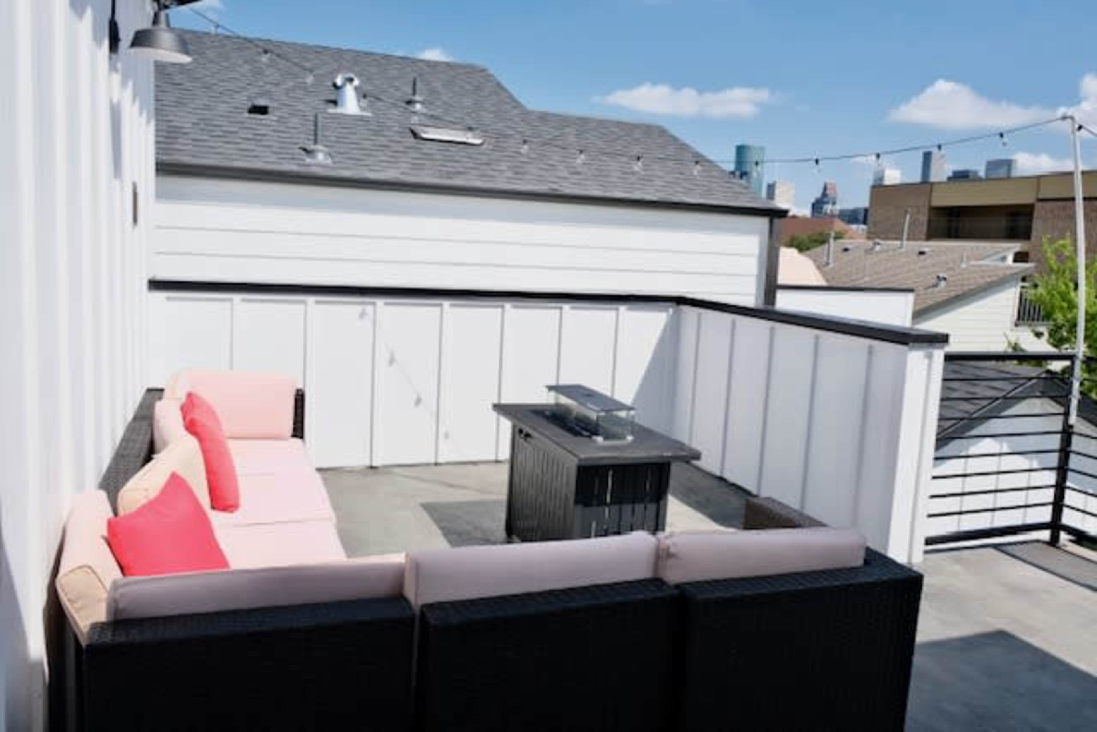 A rooftop patio features a sectional sofa with pink cushions and a black fire pit surrounded by a white railing and views of the city skyline.