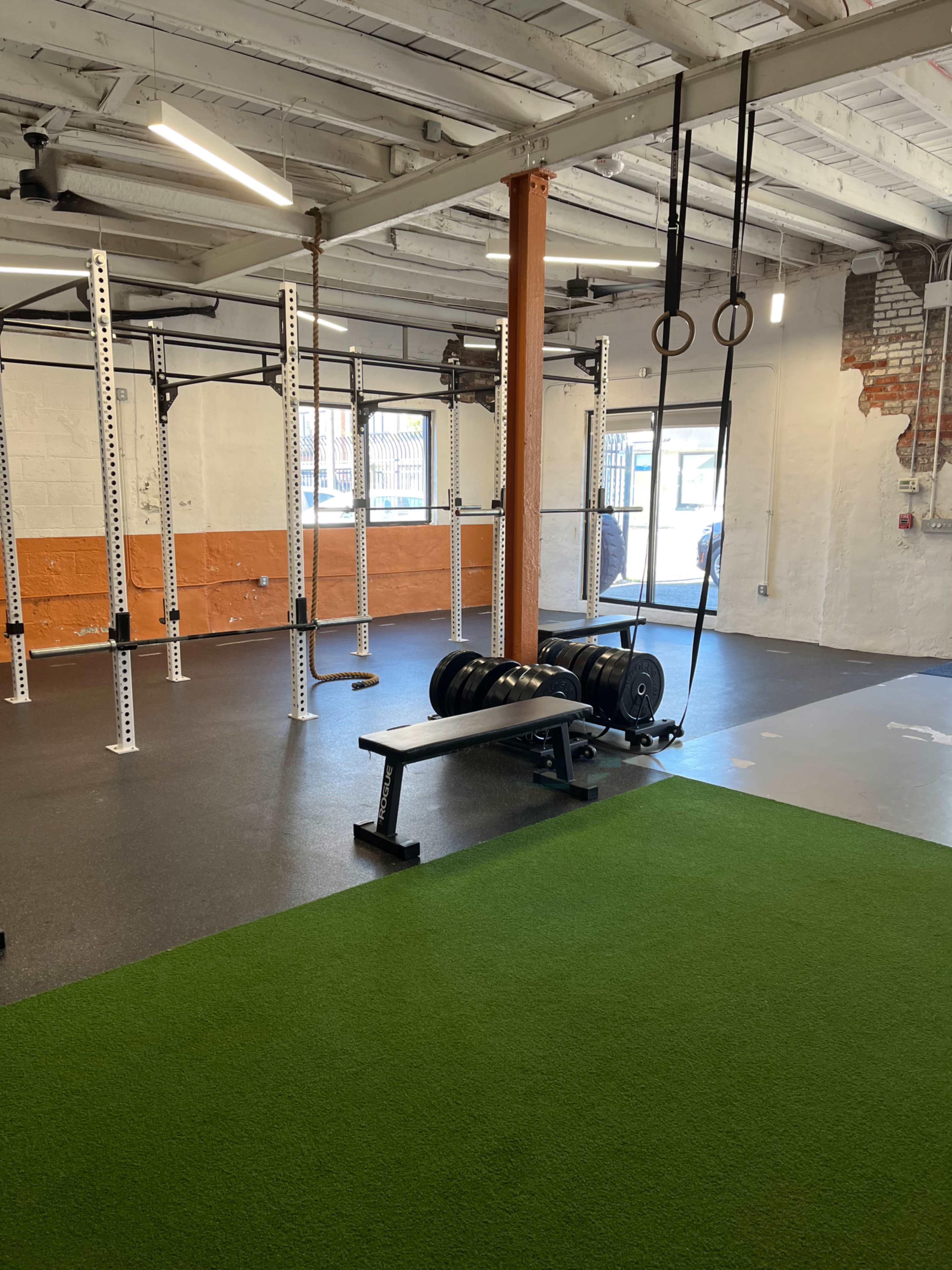 A fitness gym featuring a weight bench with plates, a green turf area, and exercise rings hanging from a ceiling beam.